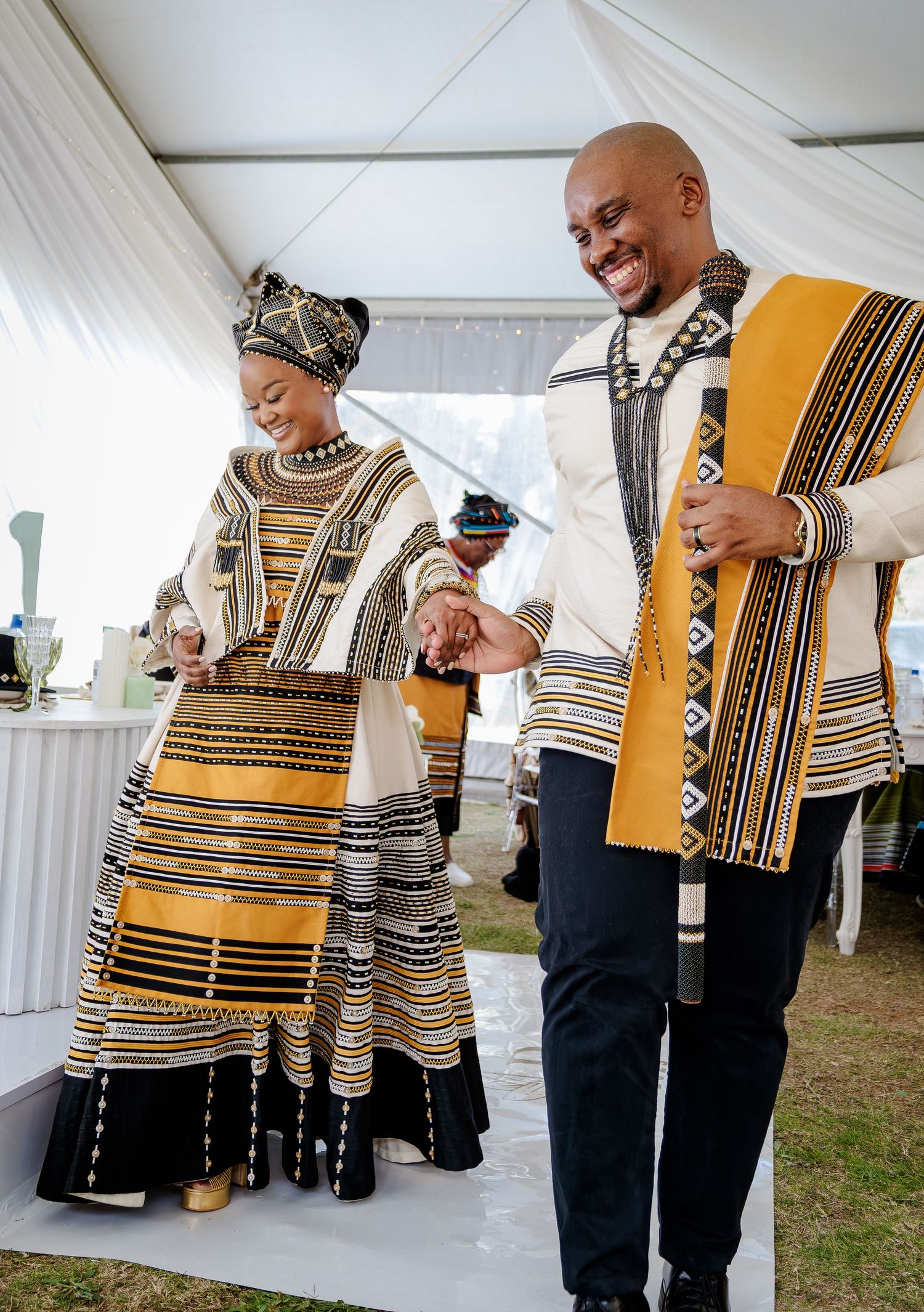 Couple in traditional Xhosa wedding attire with gold and black patterns dancing joyfully under a tent.