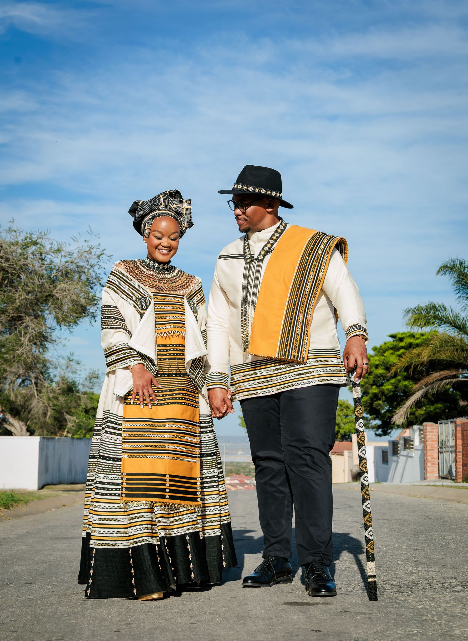 Couple walking hand-in-hand in matching traditional Xhosa wedding outfits with gold and black geometric patterns.