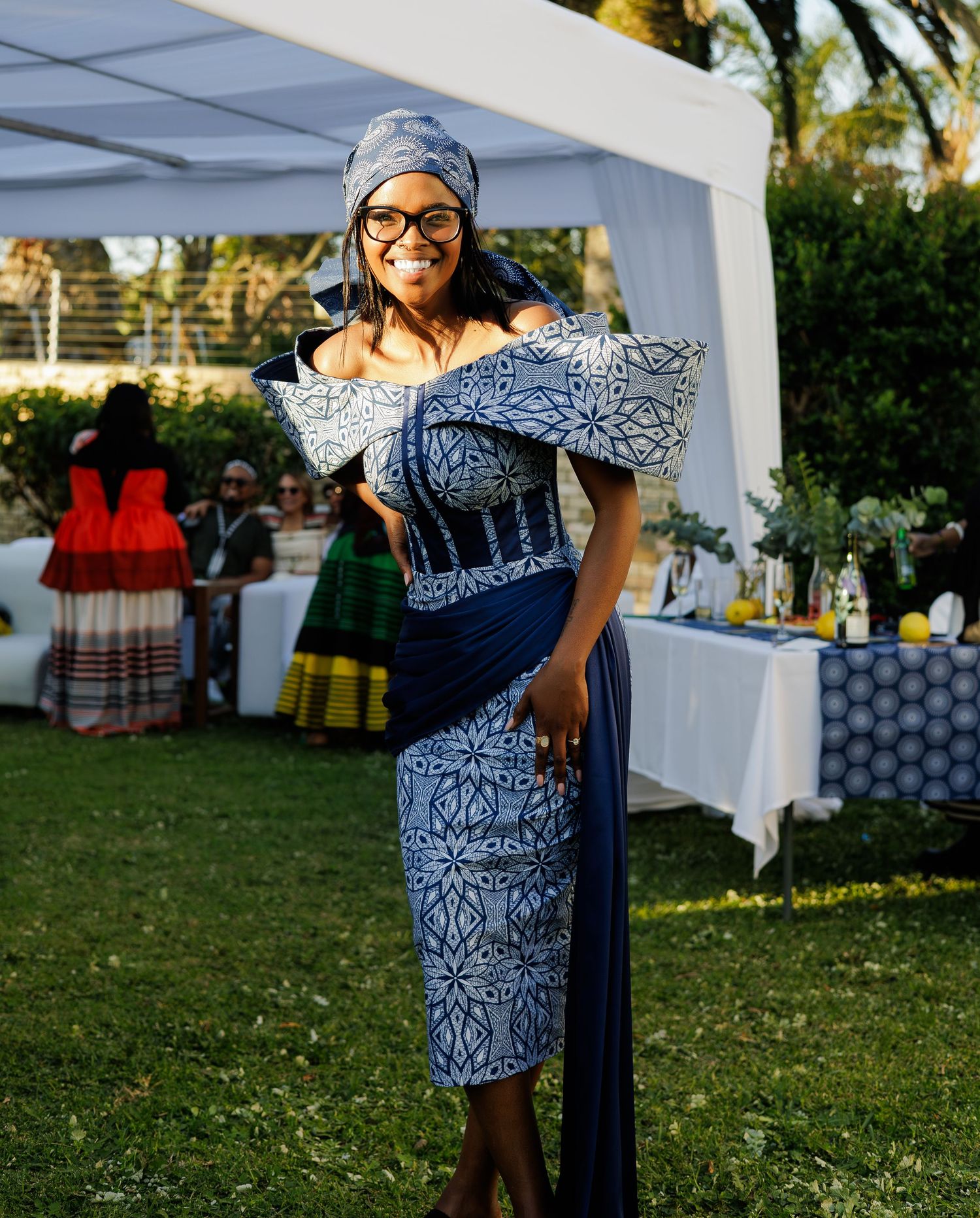 Woman in elegant blue and white Ndebele traditional dress and headwrap smiling at outdoor celebration.