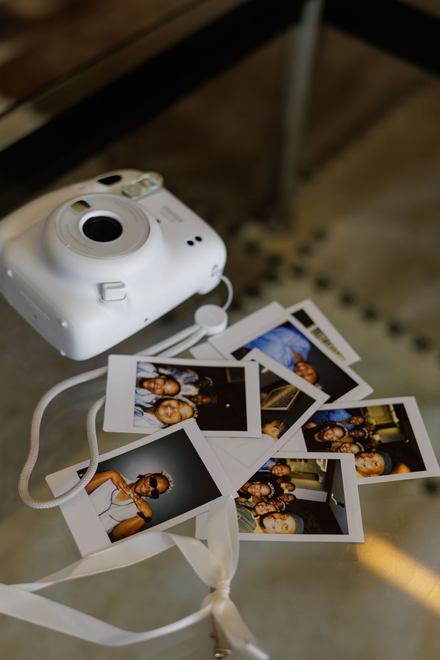 White Instax Mini camera surrounded by instant photos of wedding guests on glass table.