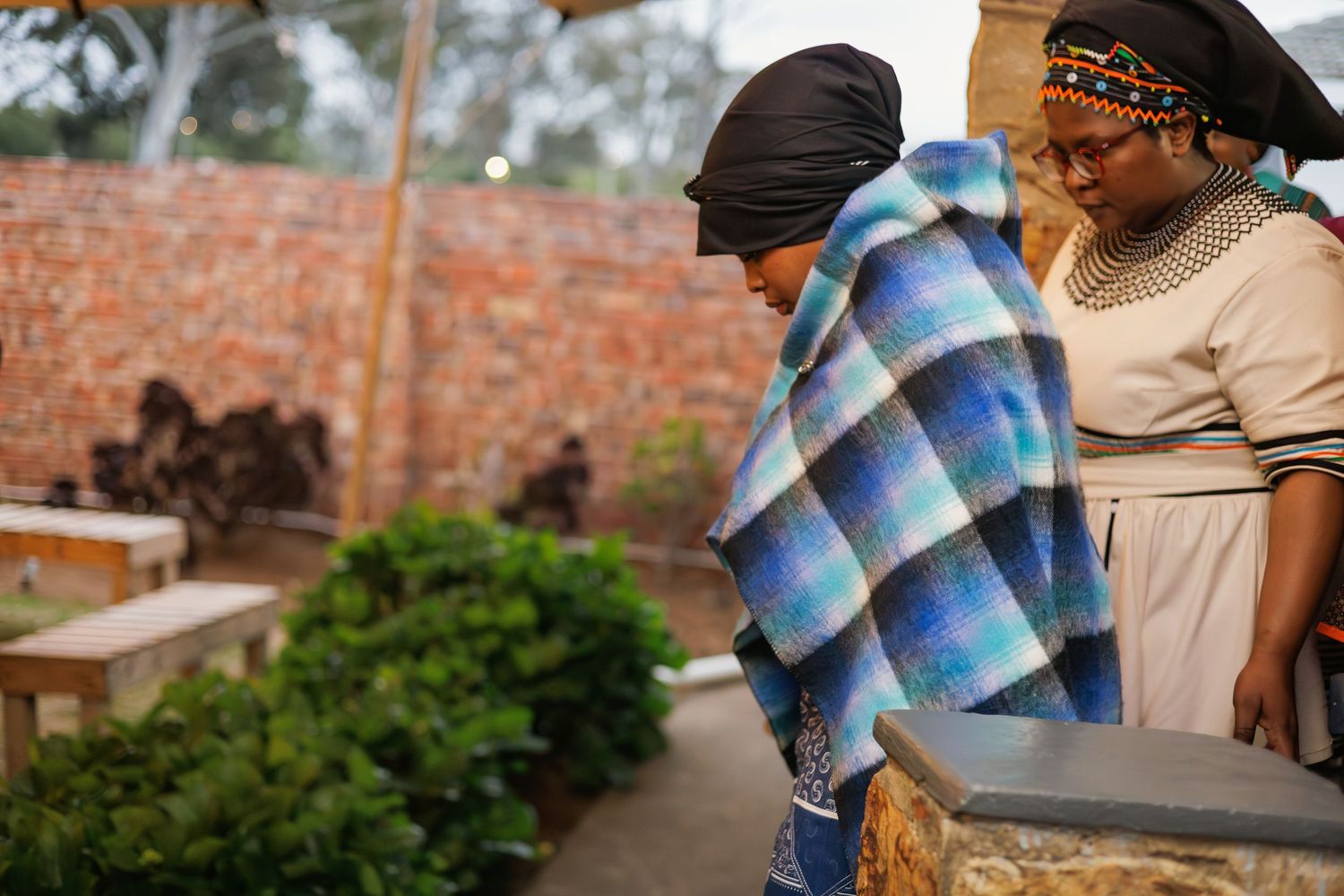 Two women in traditional African attire sharing a moment outdoors wrapped in a blue blanket.