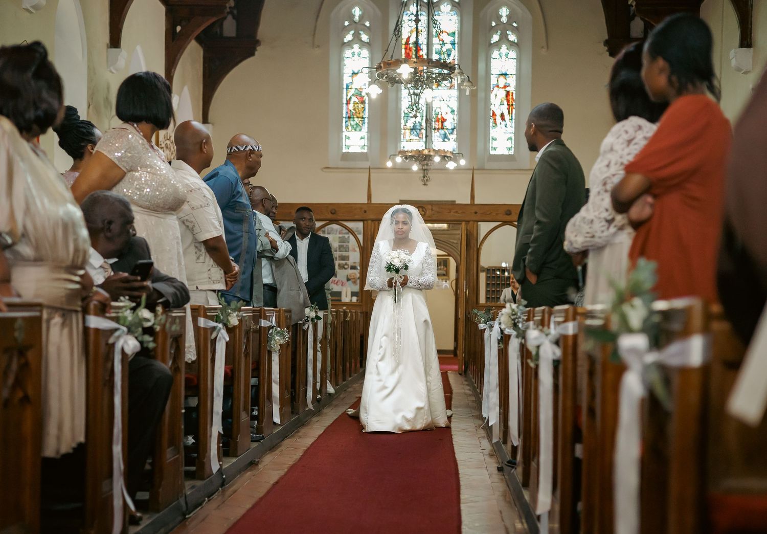 Bride in white gown and veil walks down red aisle in sunlit church.