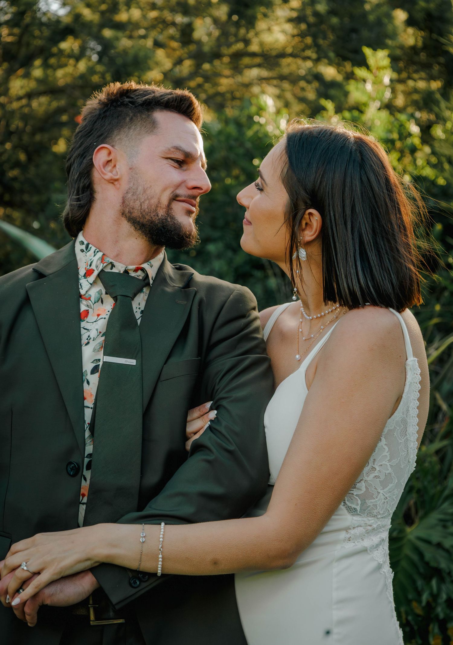 Bride and groom gaze lovingly at each other in warm sunset light among green foliage.