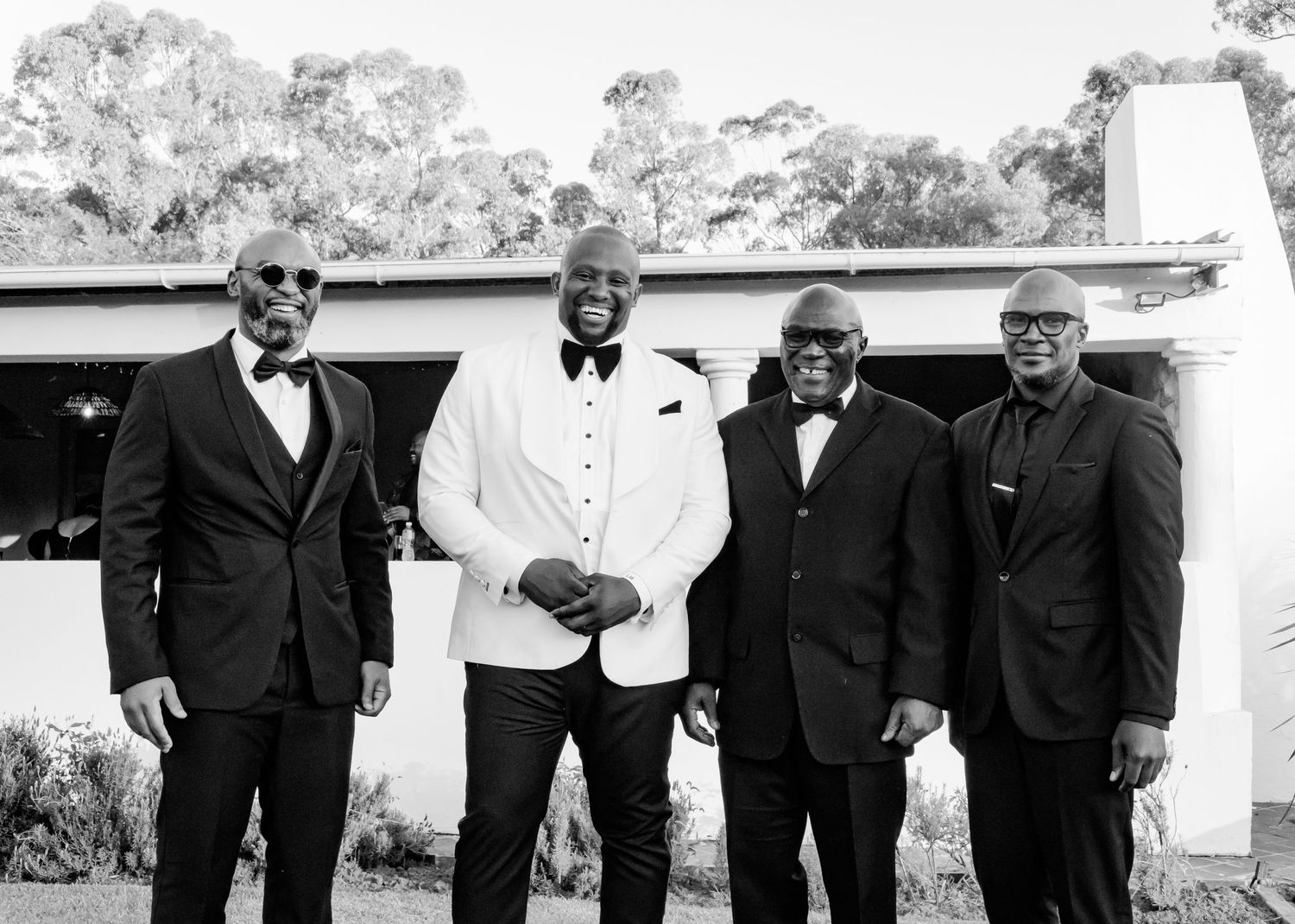 Groom in white tuxedo poses with three groomsmen in black suits outside a white wedding venue.