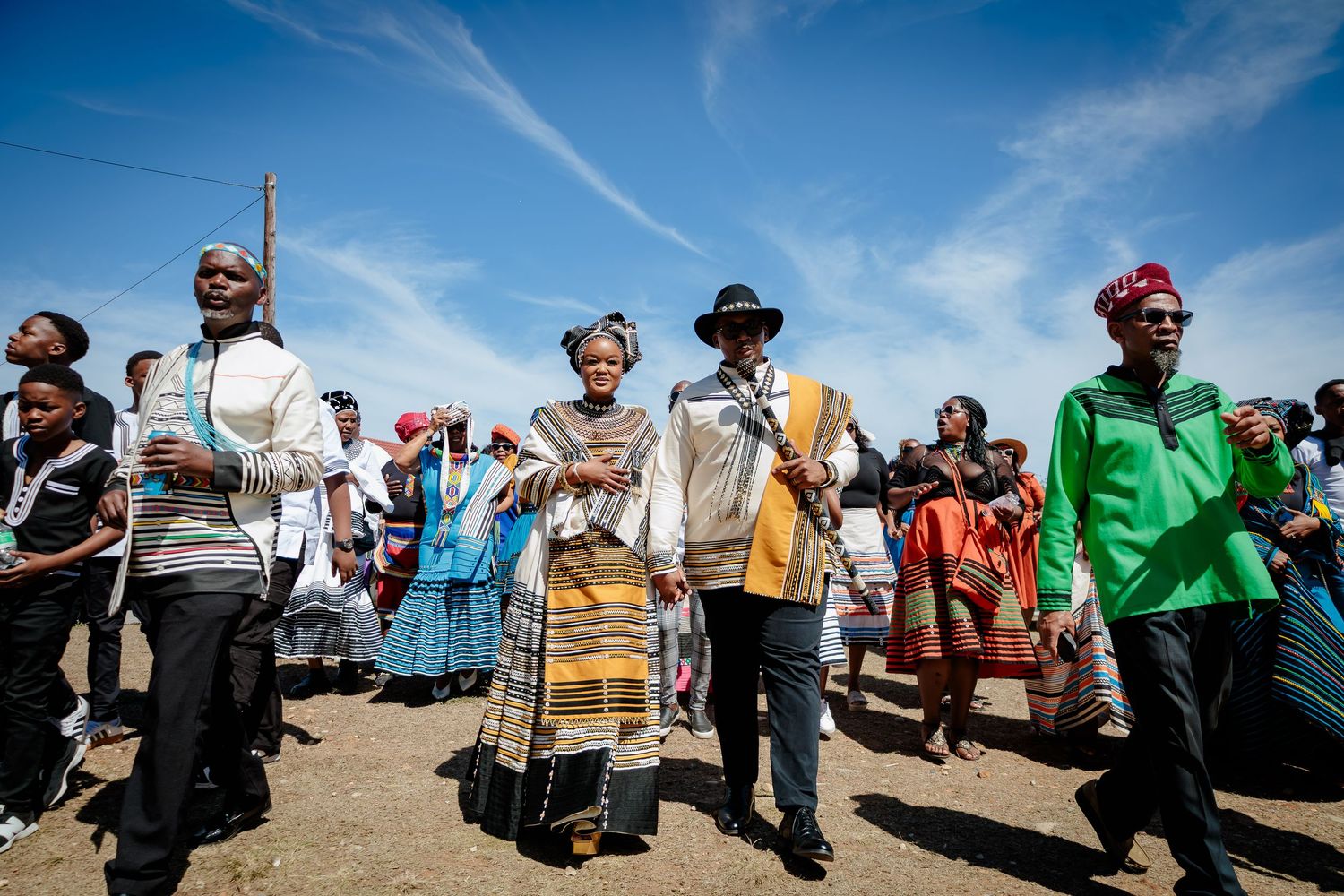 Wedding procession with couple and guests in traditional Xhosa attire walking under a blue sky.