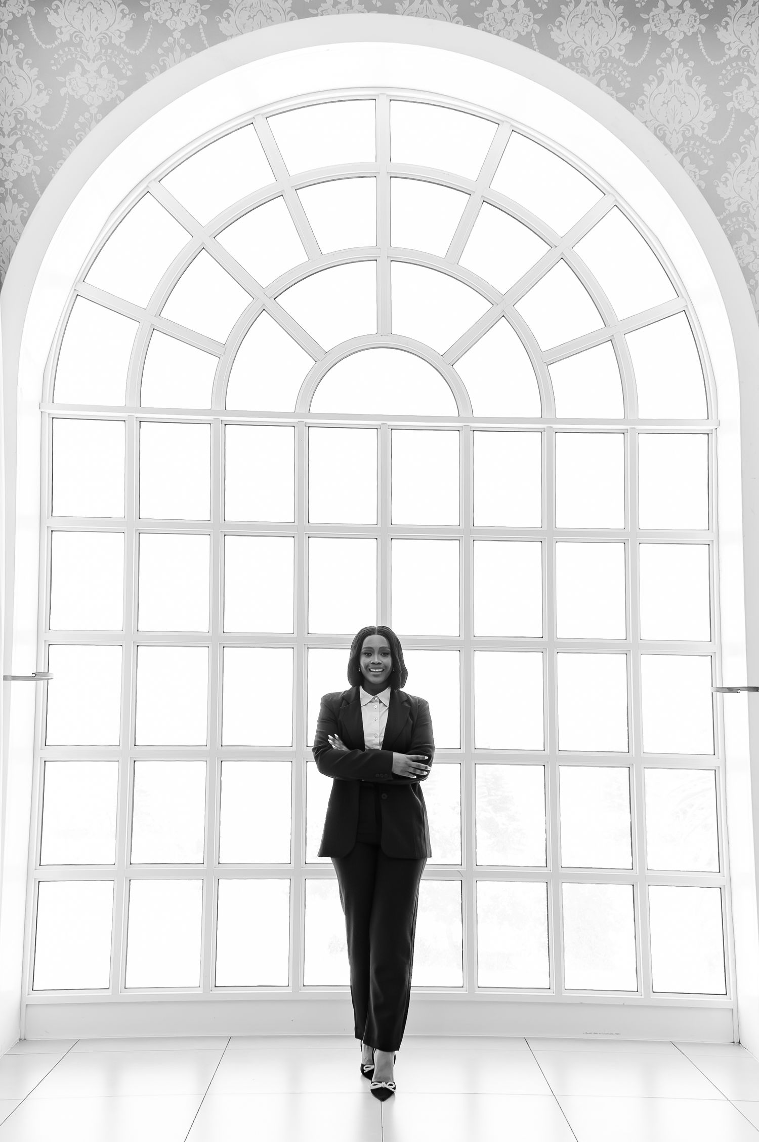 Professional woman in black suit stands confidently before large arched window in black and white photo.