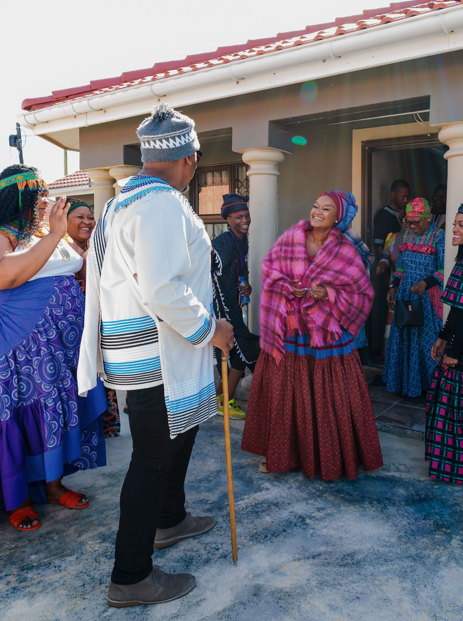 Man with walking stick in traditional Xhosa attire greets smiling woman in pink blanket at ceremony.