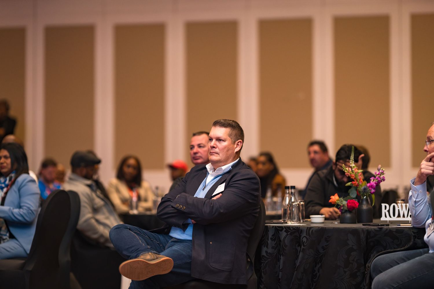 Man in blue blazer sits with arms crossed at a corporate conference event.