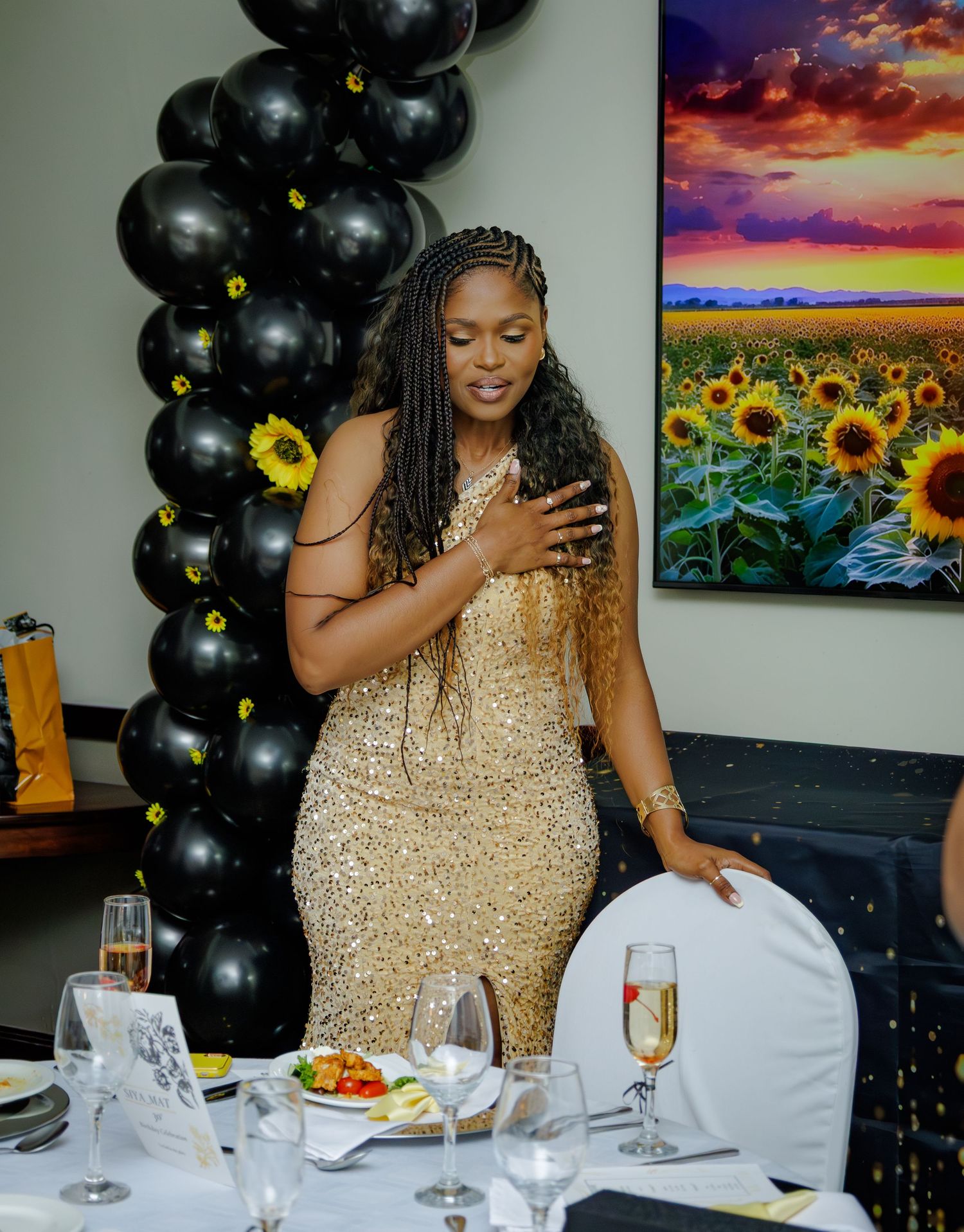 Woman in gold sequin dress posing by decorated table with black balloon column and sunflower artwork.