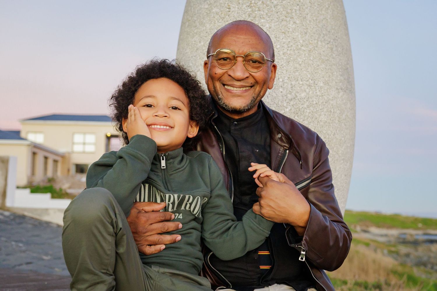 An elderly Black man smiles warmly while hugging a young curly-haired boy outdoors near a coastal monument.