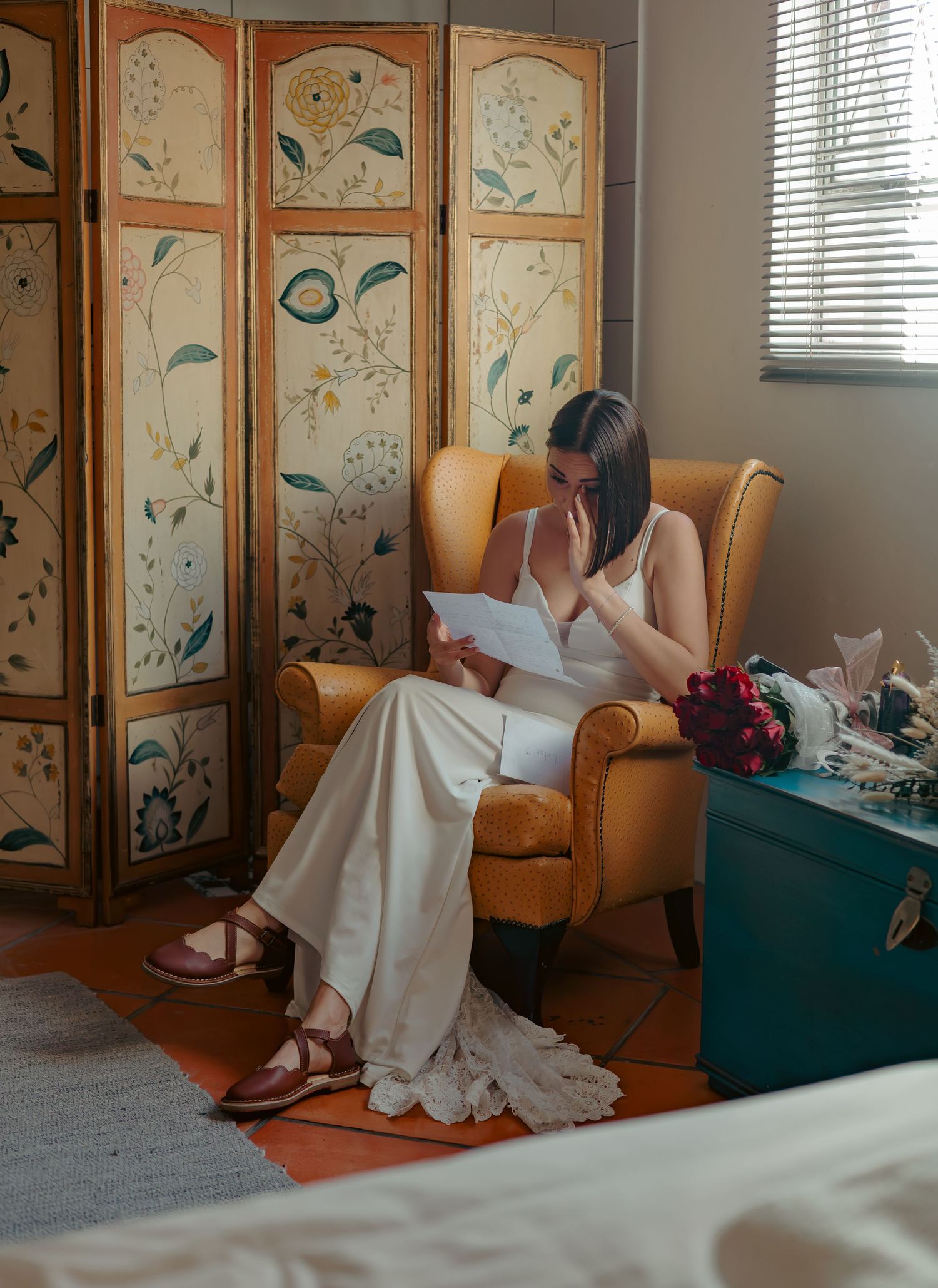 Bride in elegant white dress reading a letter seated in yellow chair by decorative screen.