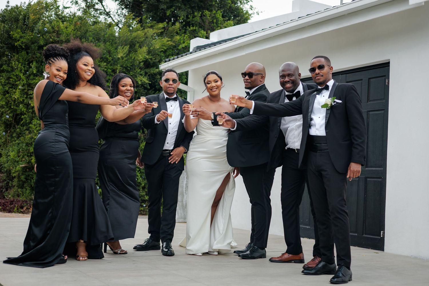 Wedding party in black attire celebrates with bride in white gown outside white building.