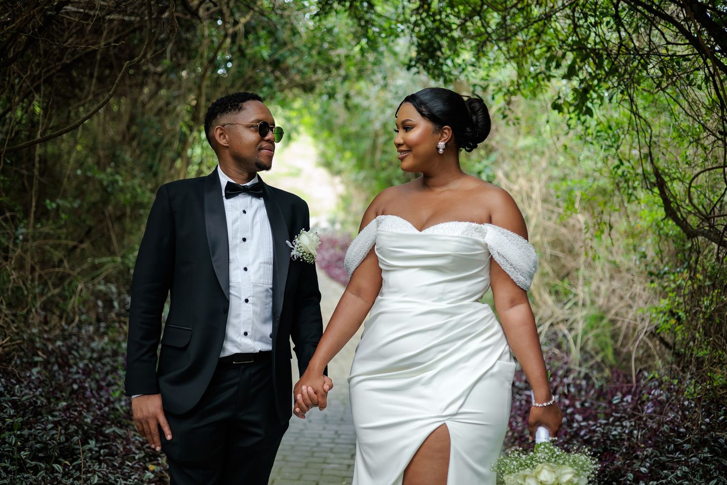Newlywed couple in wedding attire share a loving glance while walking through green garden.