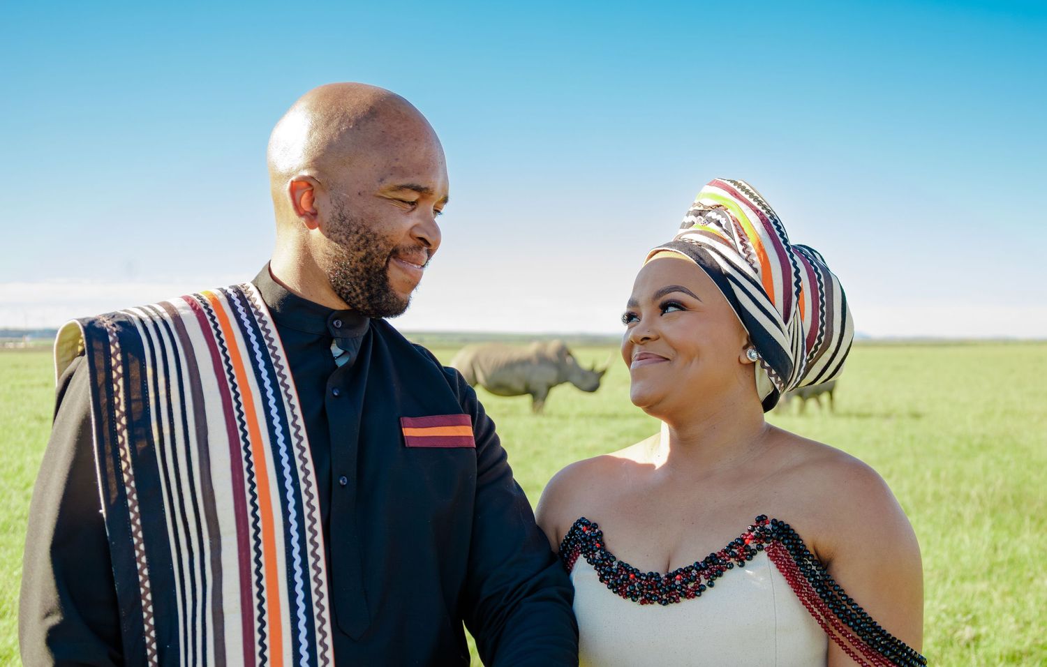 Couple in traditional African attire smiling at each other in a green field with a rhino in the background.