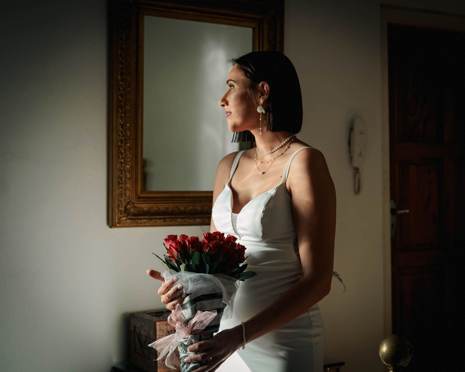 Elegant bride in white slip dress holding red roses bouquet while gazing out window at mirror.
