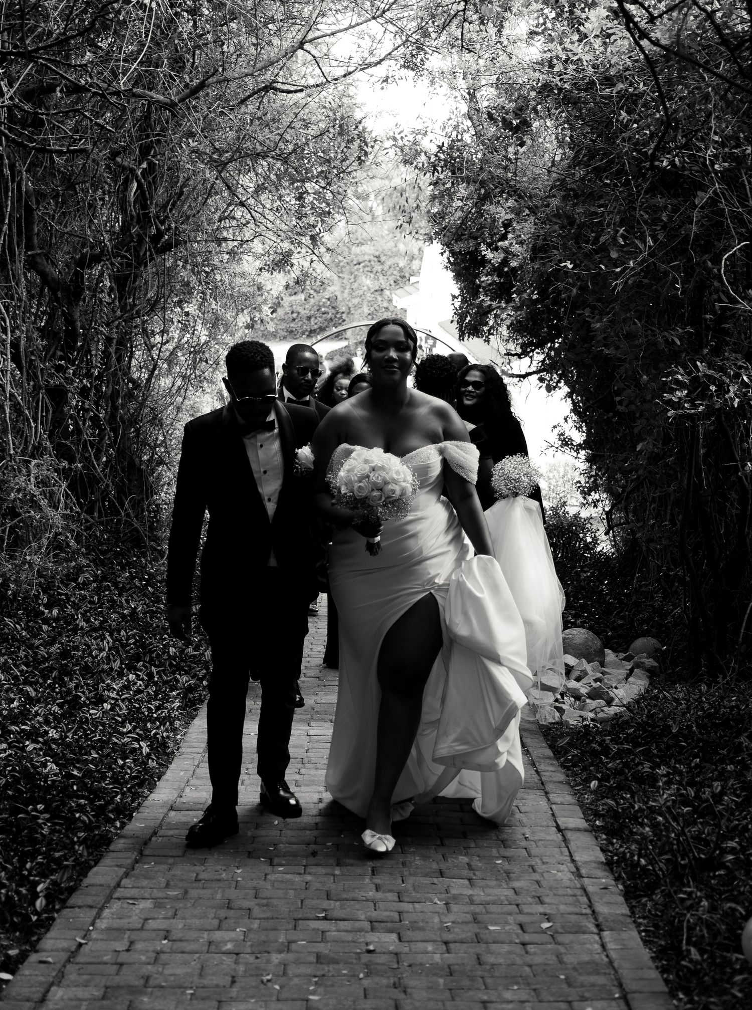 Black and white photo of bride and groom leading wedding party down tree-lined path.