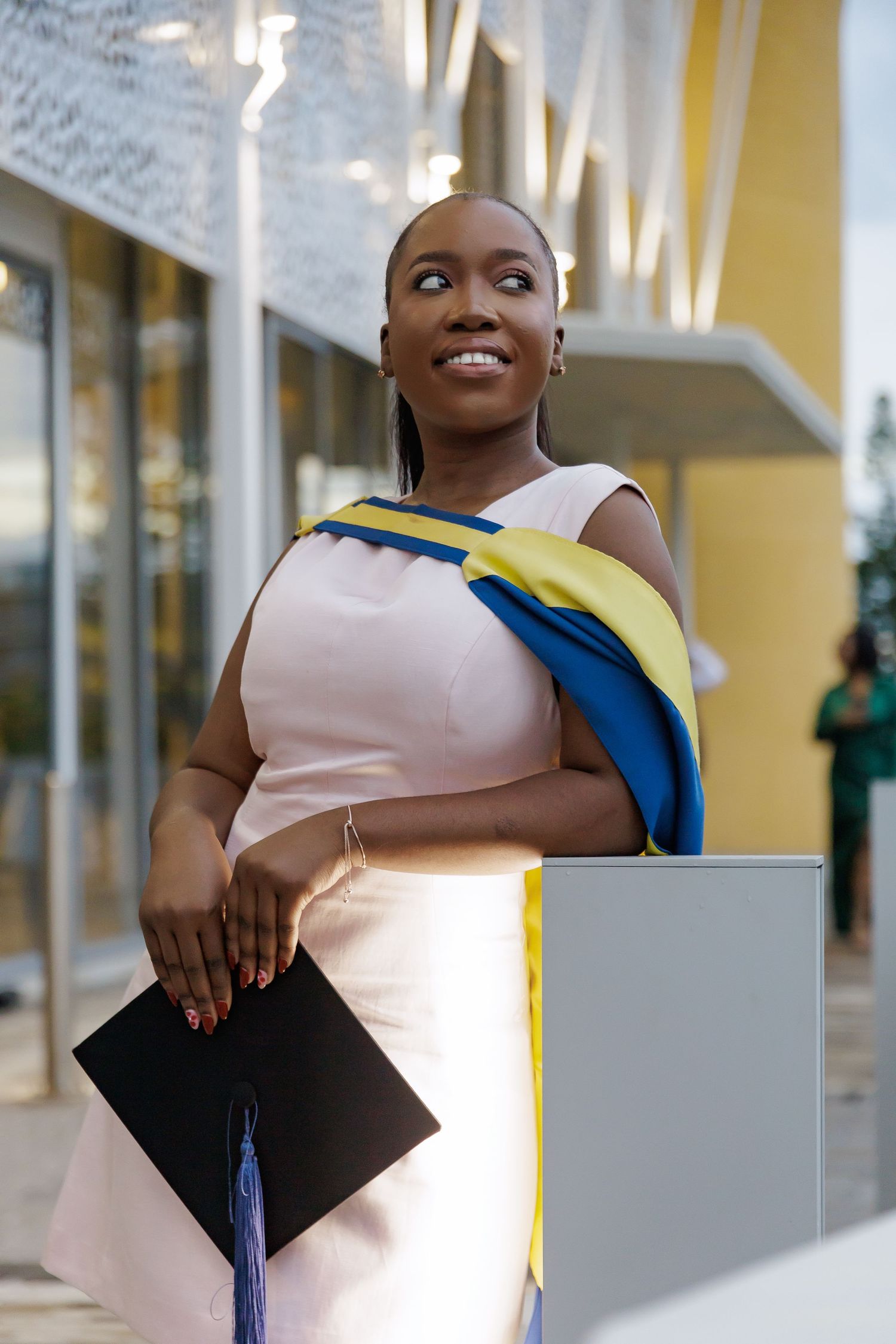 A Black female graduate in a pink dress holds her mortarboard while wearing blue and yellow academic hood.