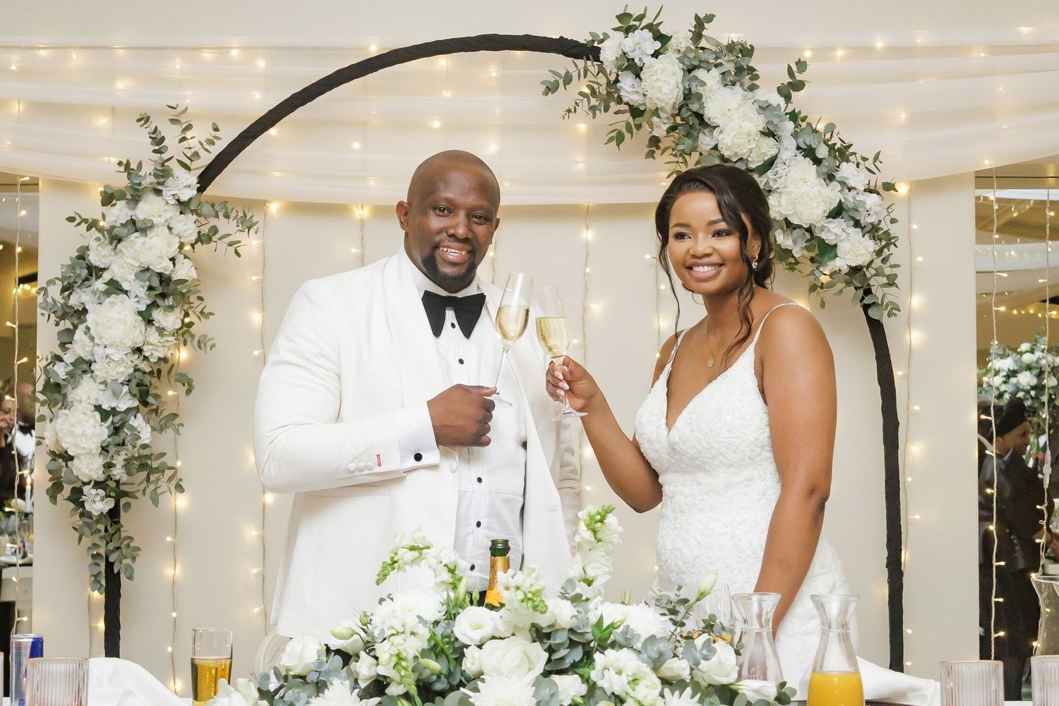 Bride and groom toast with champagne under a floral arch at their white and green wedding reception.