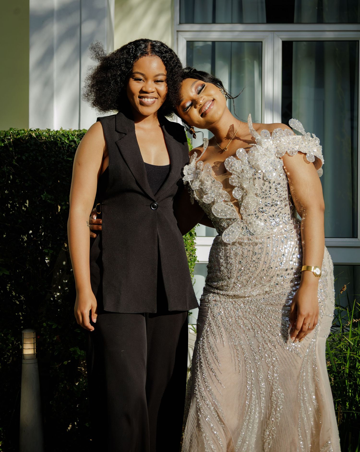 Two women posing outdoors, one in black pantsuit and one in silver beaded formal gown.