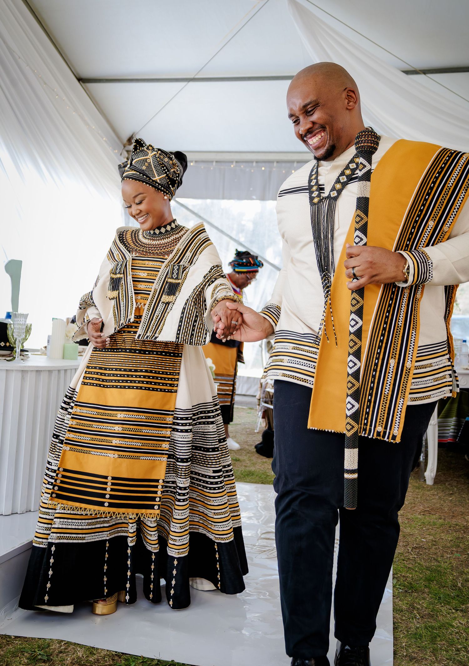 Smiling couple in matching traditional Xhosa black and gold wedding attire walking together.