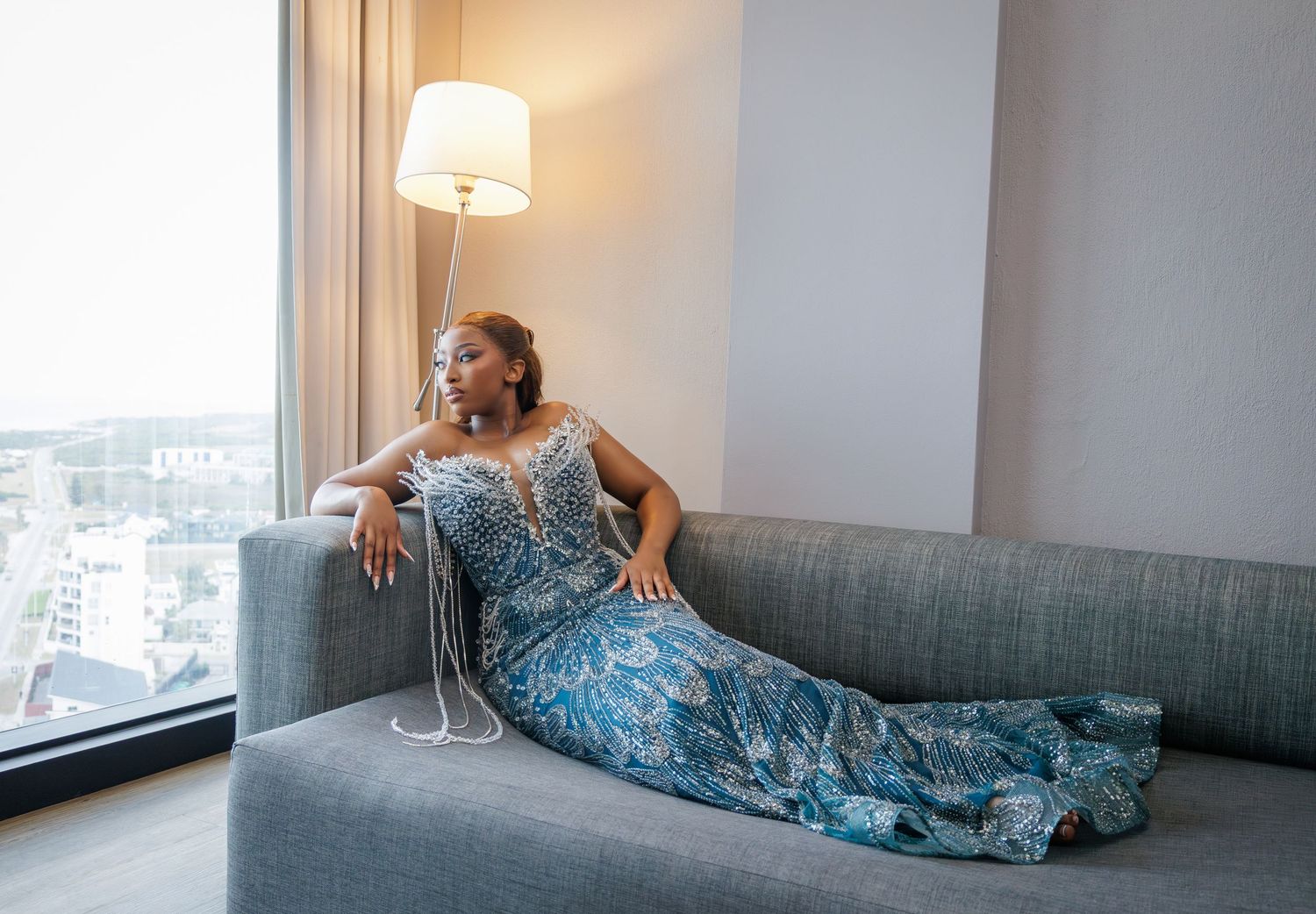 Woman in blue beaded gown reclines gracefully on grey sofa near window with city view.