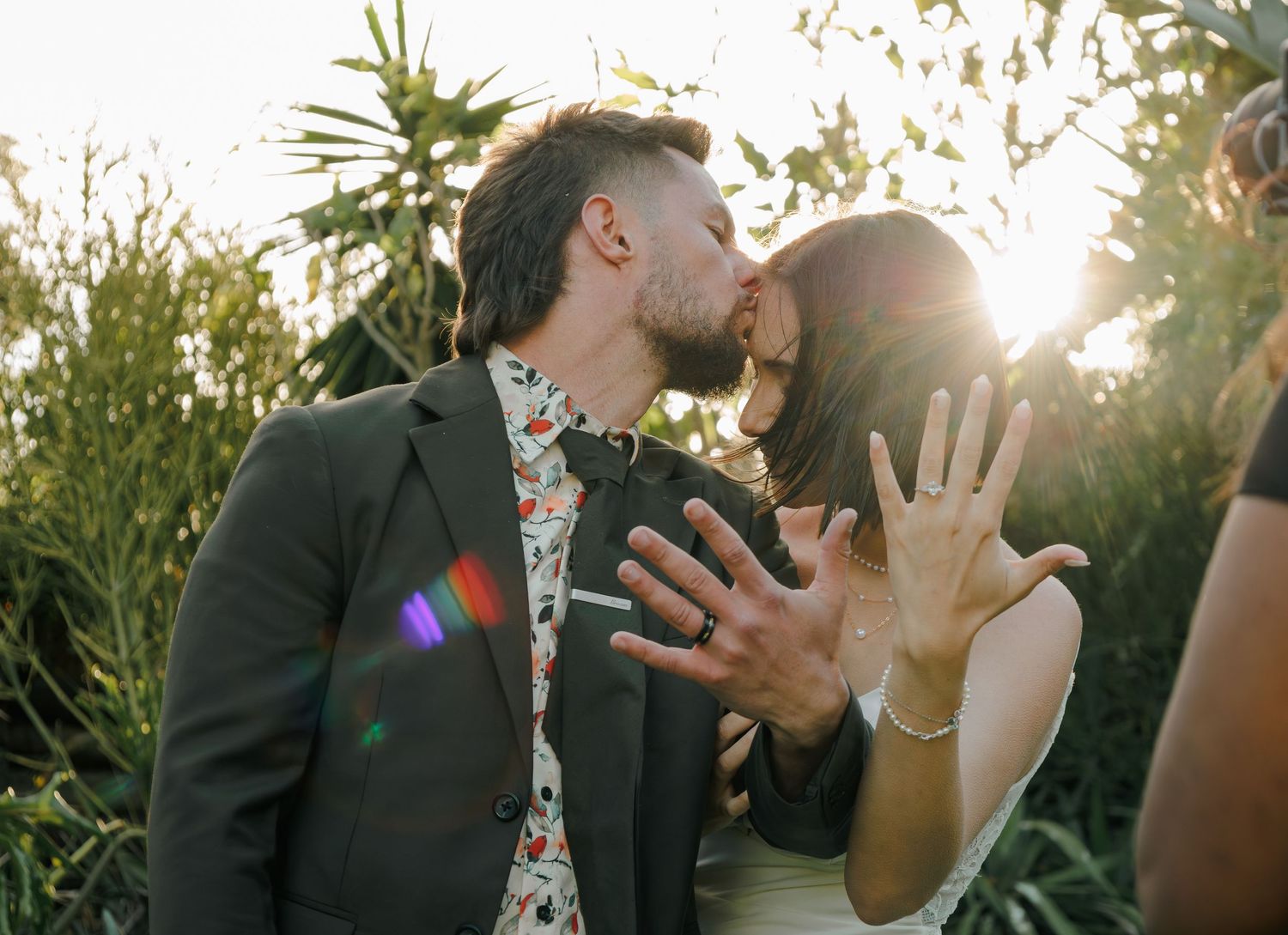 Newlyweds kissing and showing wedding rings in sunlit garden ceremony.