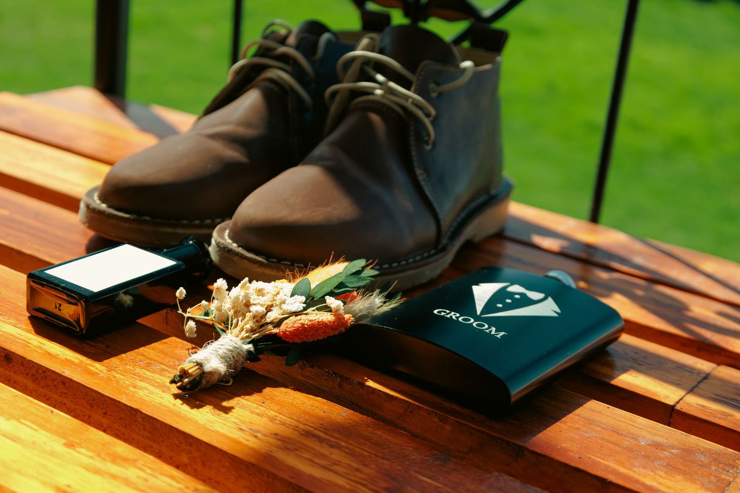 Groom wedding details flat lay with brown leather boots, boutonniere, and groom wallet on wood.