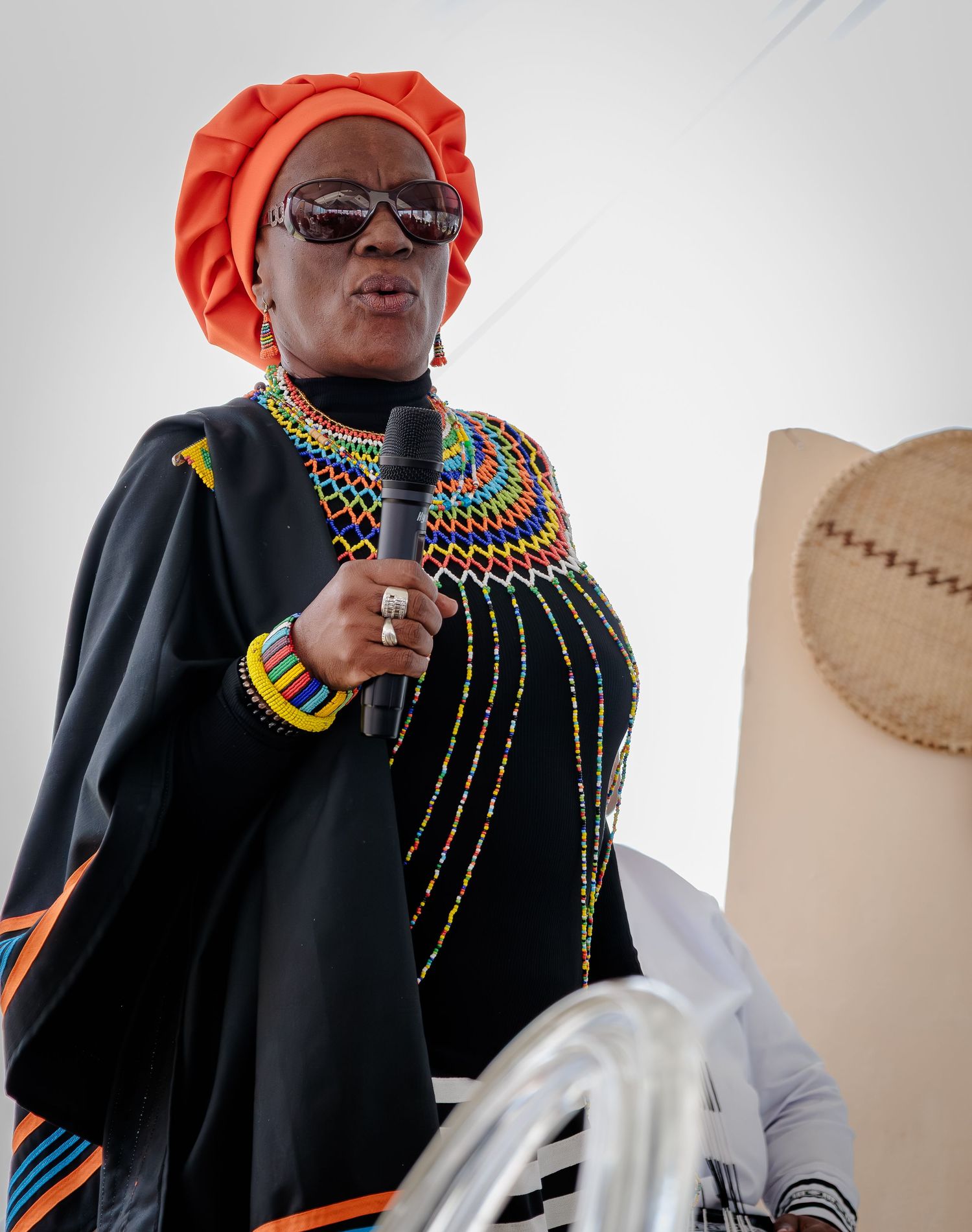 Woman in traditional Zulu beaded attire and orange headwrap speaking into a microphone.