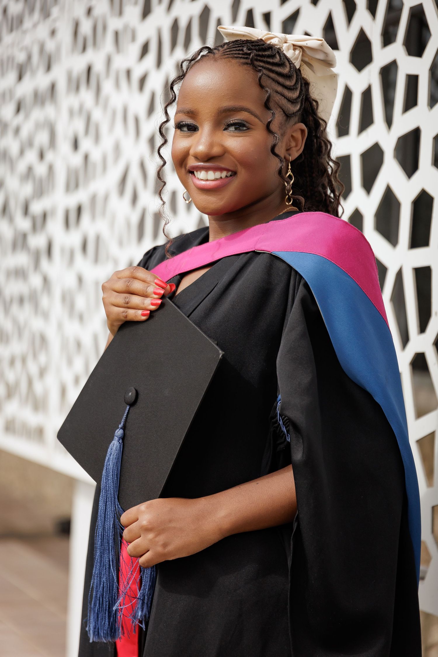 A smiling Black female graduate in black gown with pink and blue hood holds her mortarboard outdoors.