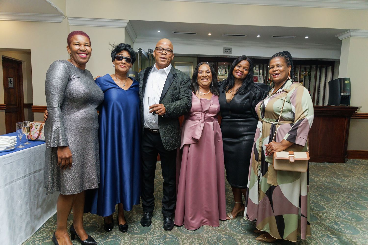 Group of six people posing together and smiling at a formal indoor celebration.