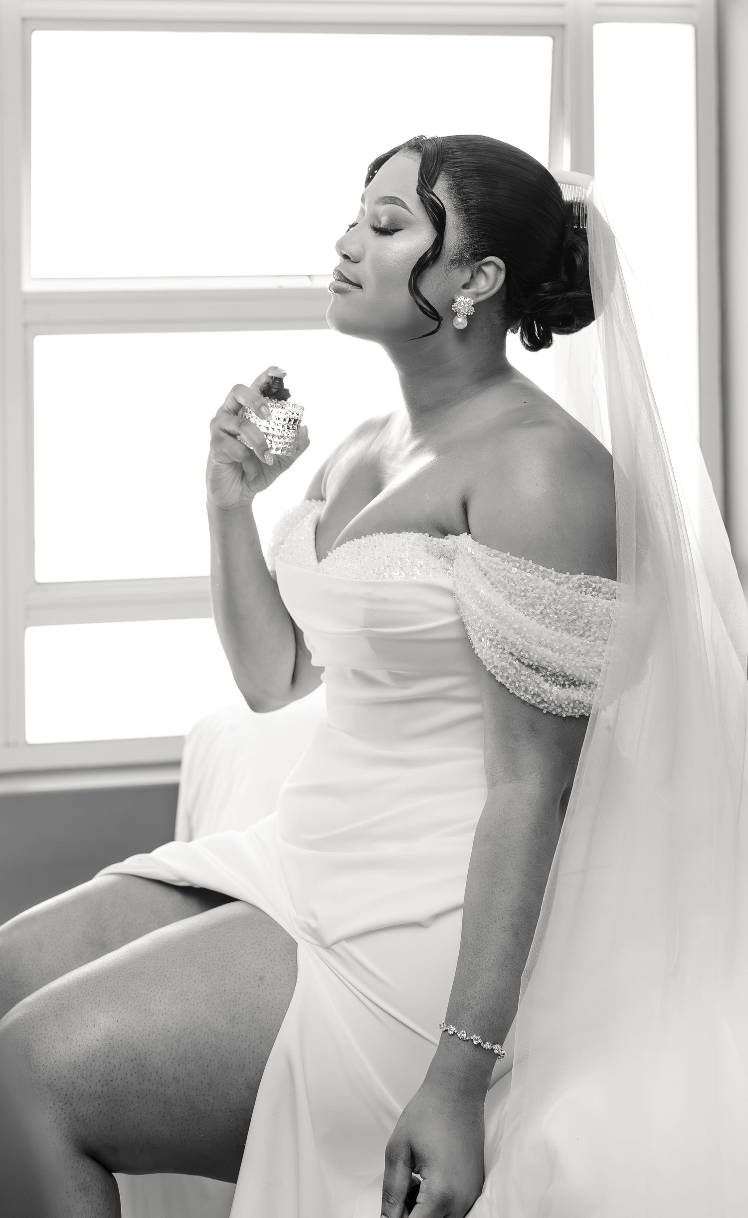 Elegant black and white portrait of bride in wedding gown applying perfume by sunlit window.