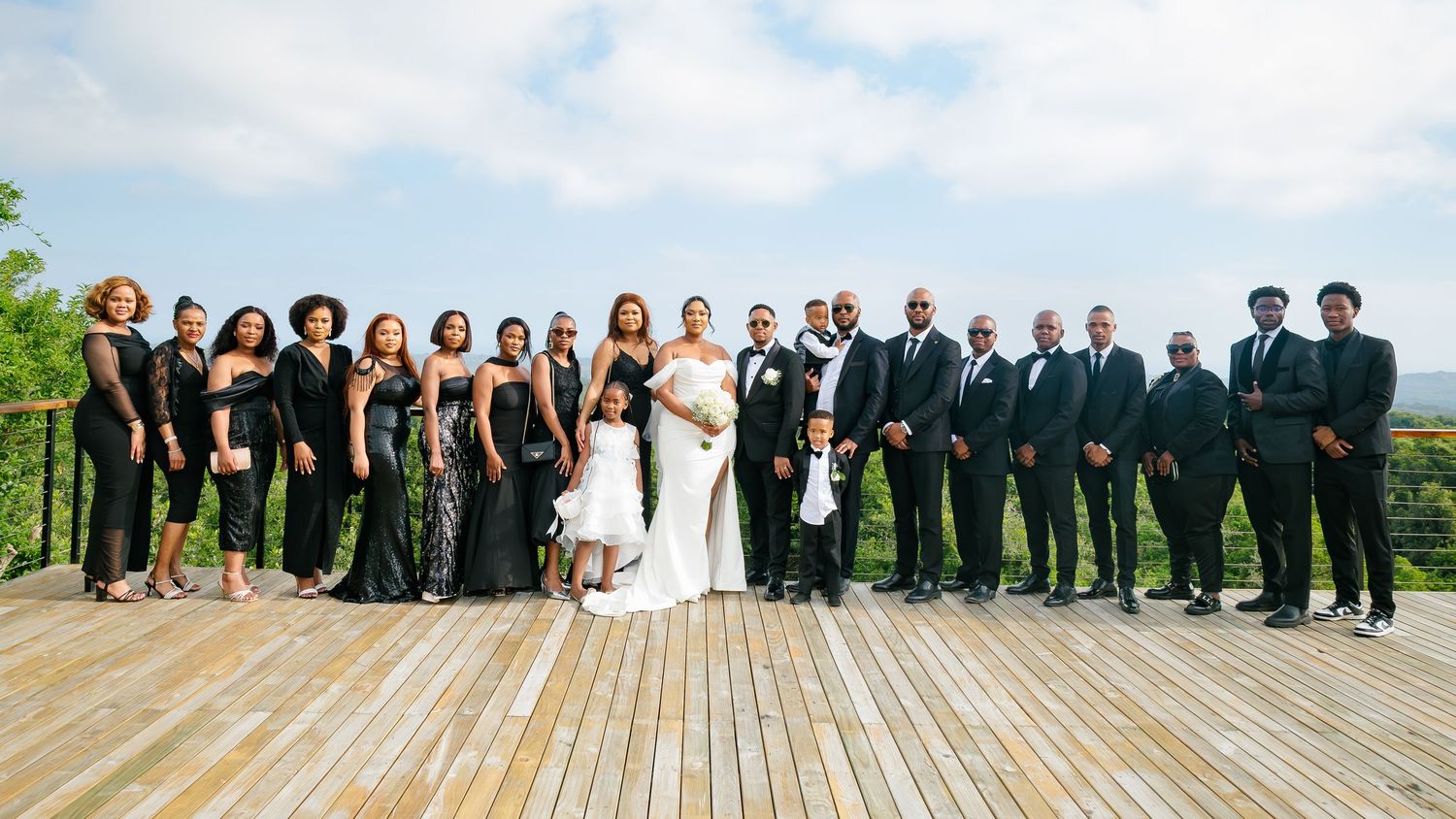 Elegant wedding party in black formal wear posing on a wooden deck with lush landscape backdrop.