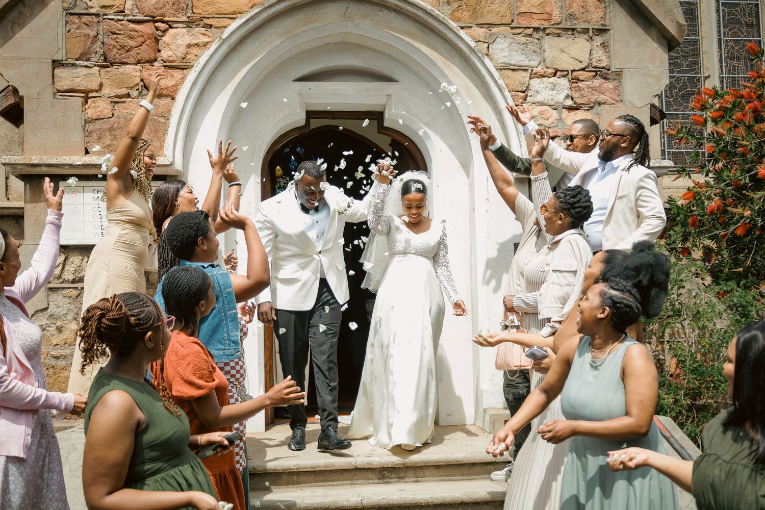 Newlyweds celebrate with confetti outside stone church entrance surrounded by cheering guests.
