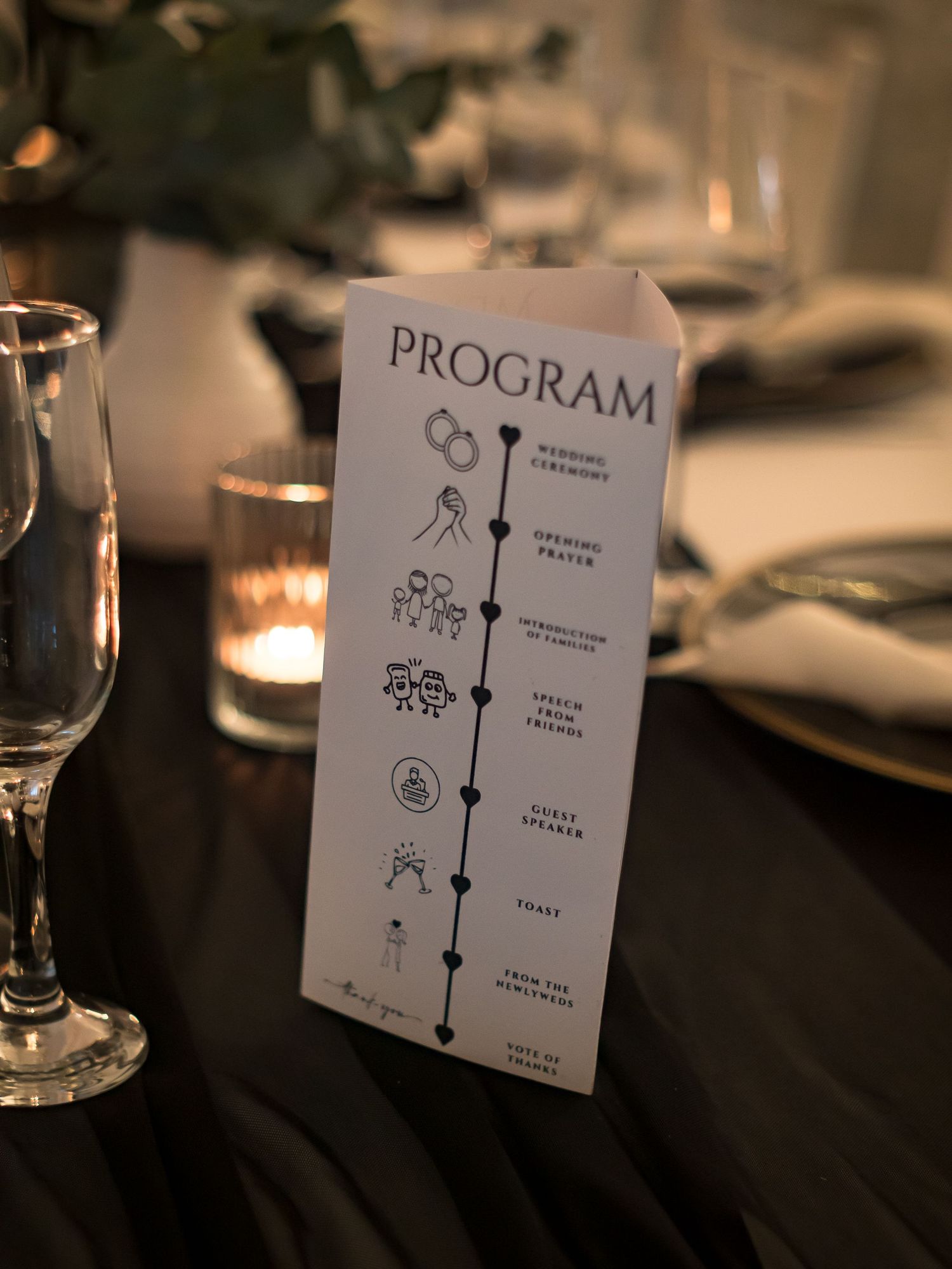 Elegant wedding program card displayed on a decorated reception table with candles and glassware.