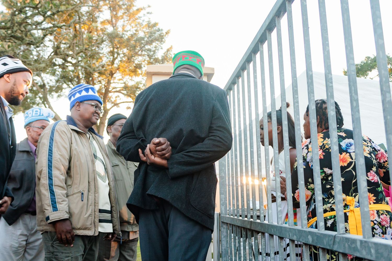 Men in traditional African caps converse through iron gate at outdoor cultural event.