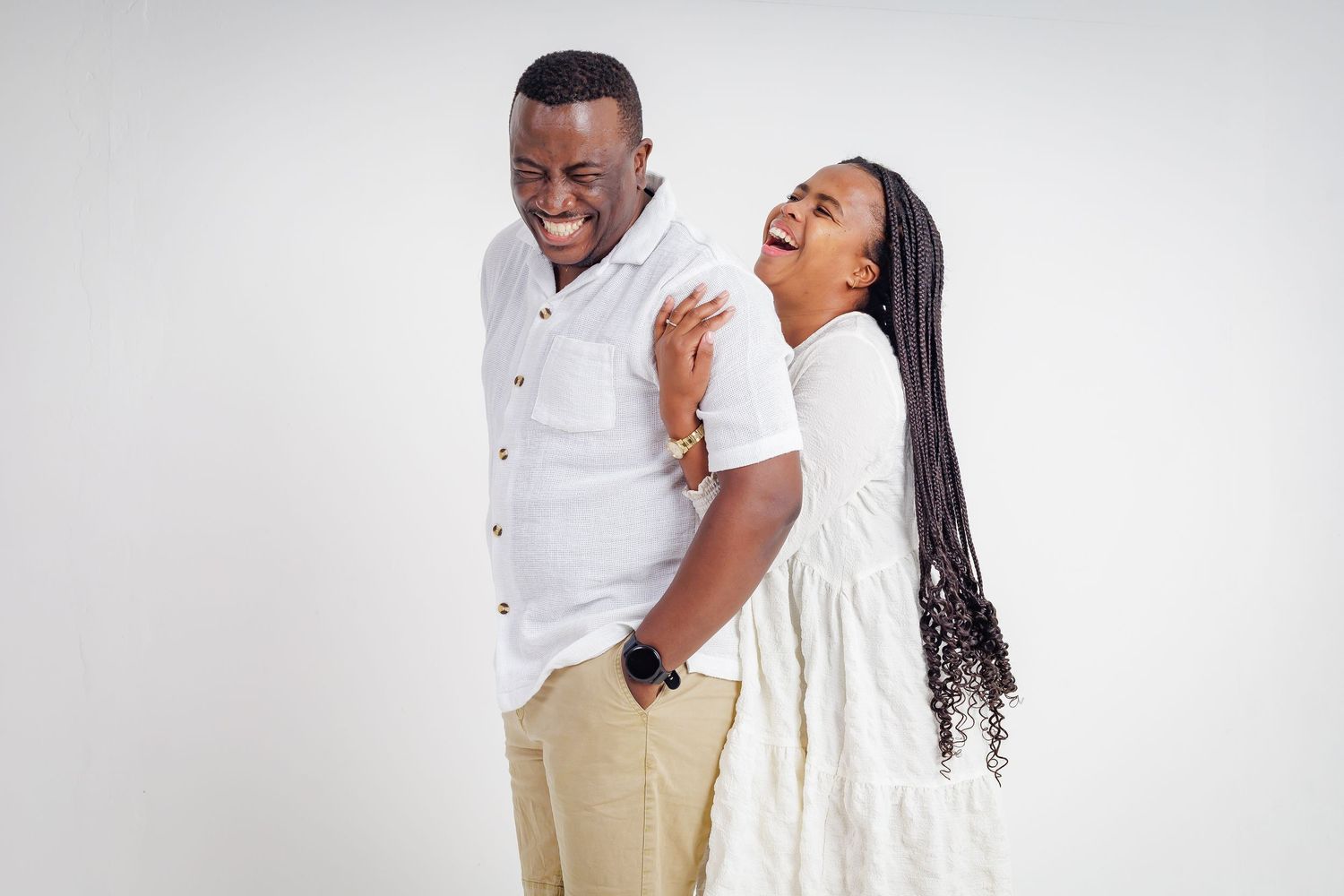 Happy couple in white outfits laughing and enjoying a playful moment in a white studio.
