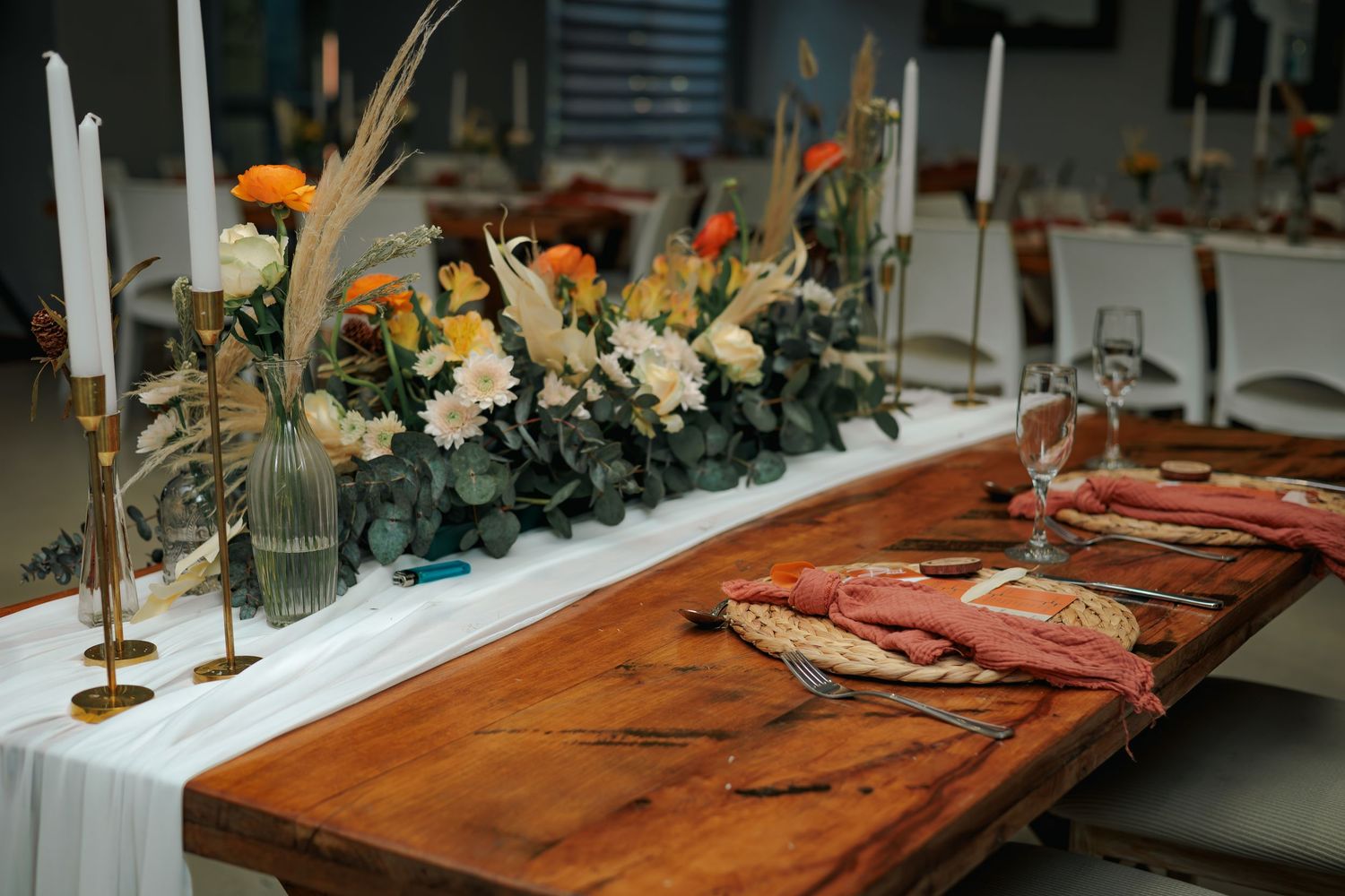 Rustic wooden head table adorned with white and orange floral arrangements, pampas grass, and tall gold candle holders.