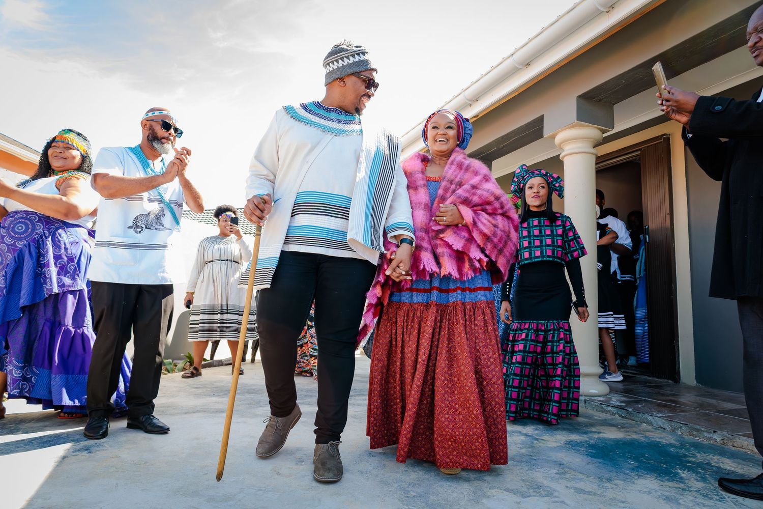 A man with a cane in traditional attire walks alongside a woman in colorful dress during a cultural ceremony.