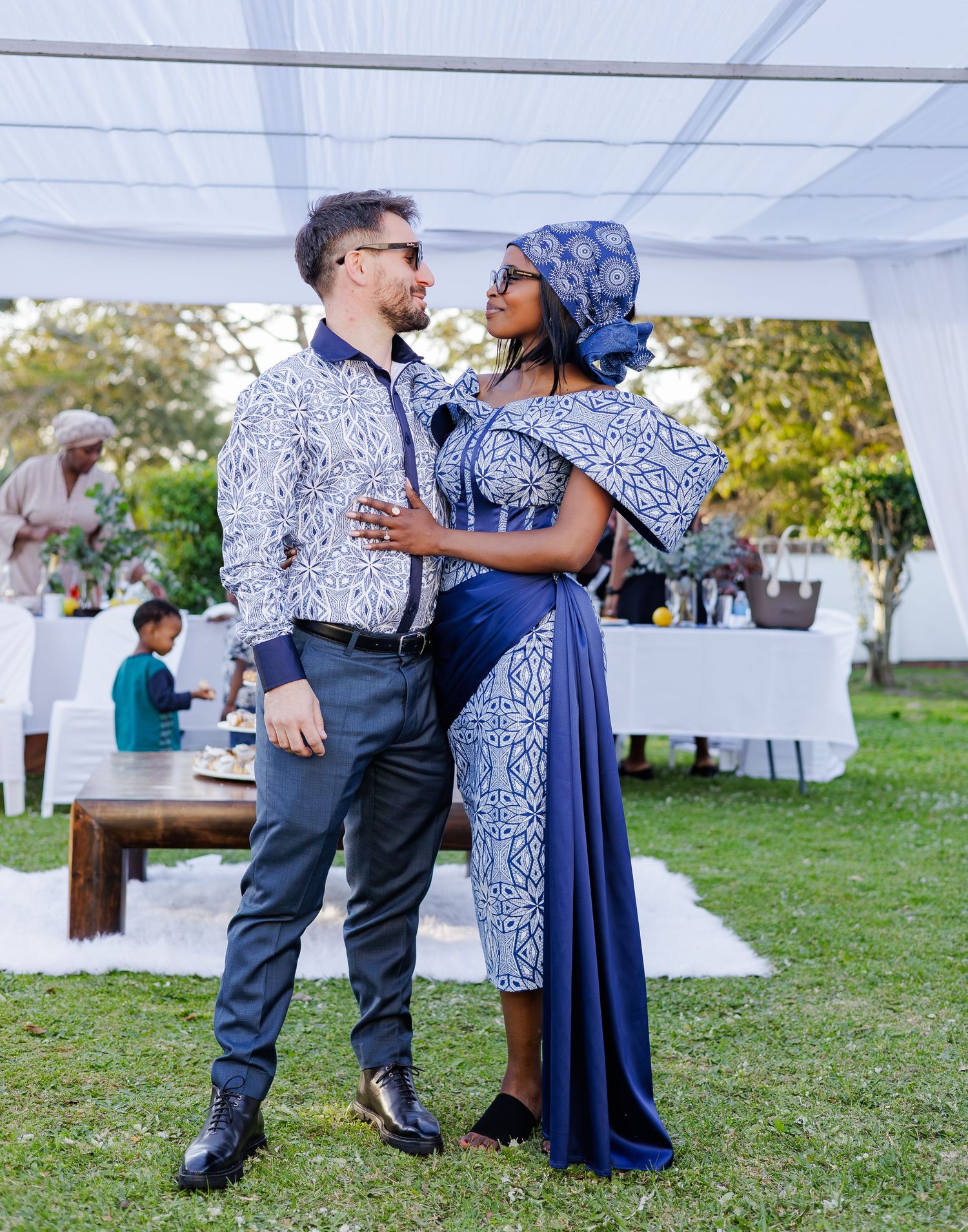 Couple in coordinating blue African print attire gazing at each other at an outdoor traditional event.