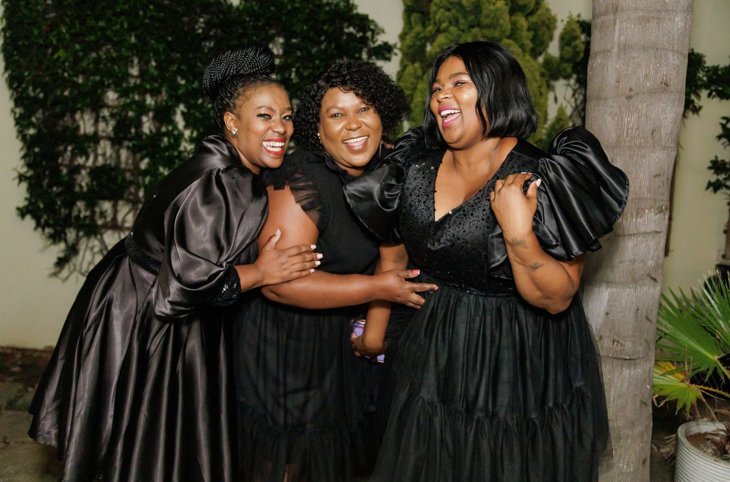 Three women in black dresses laughing and embracing outdoors at a night event.