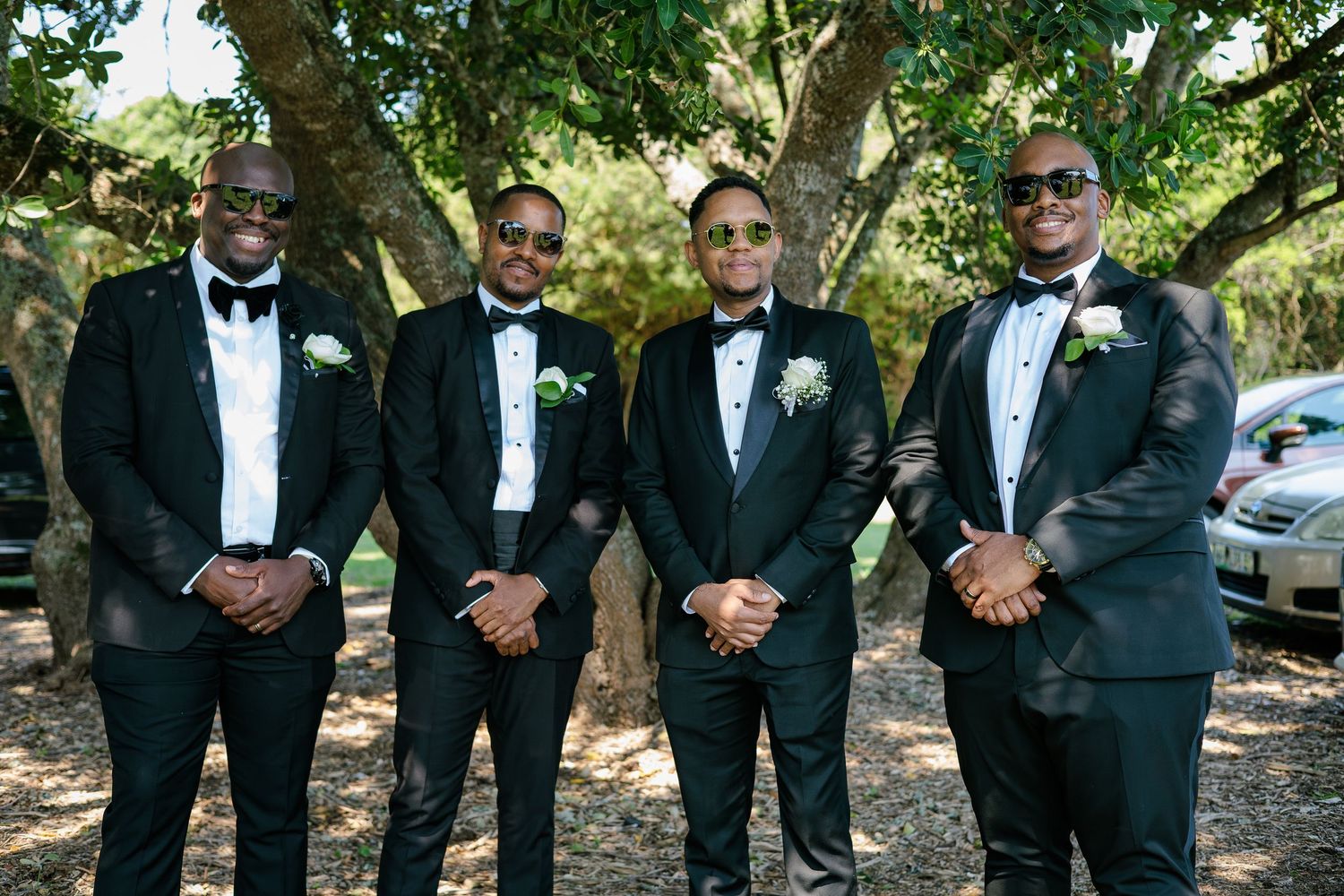 Four groomsmen in black tuxedos with bow ties and white boutonnieres posing under trees at a wedding.
