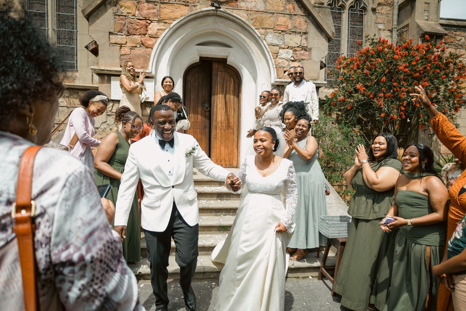 Bride and groom hold hands exiting church as bridesmaids in green dresses celebrate.