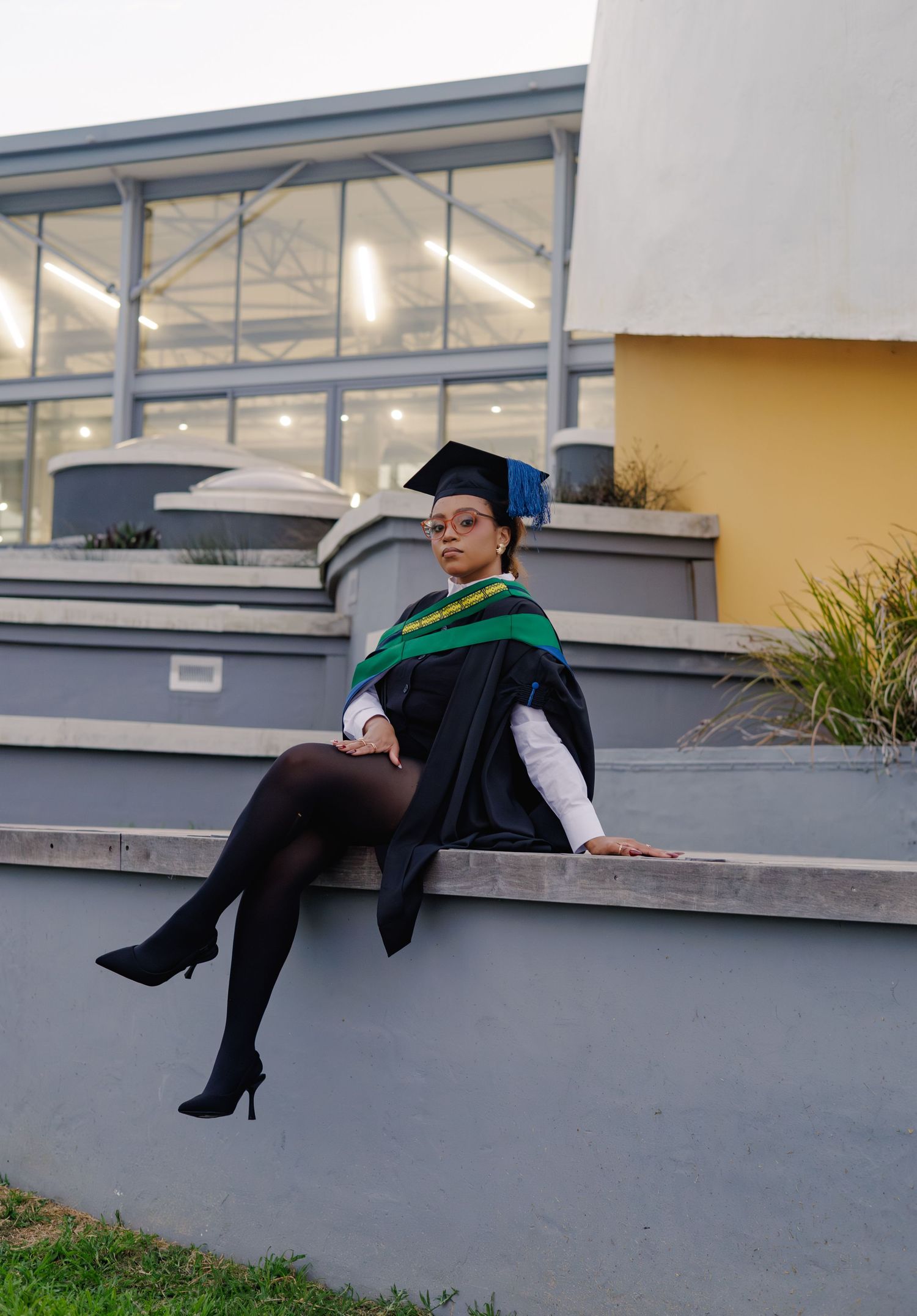 Graduate in black cap and gown with green hood sits on concrete steps outside university building.