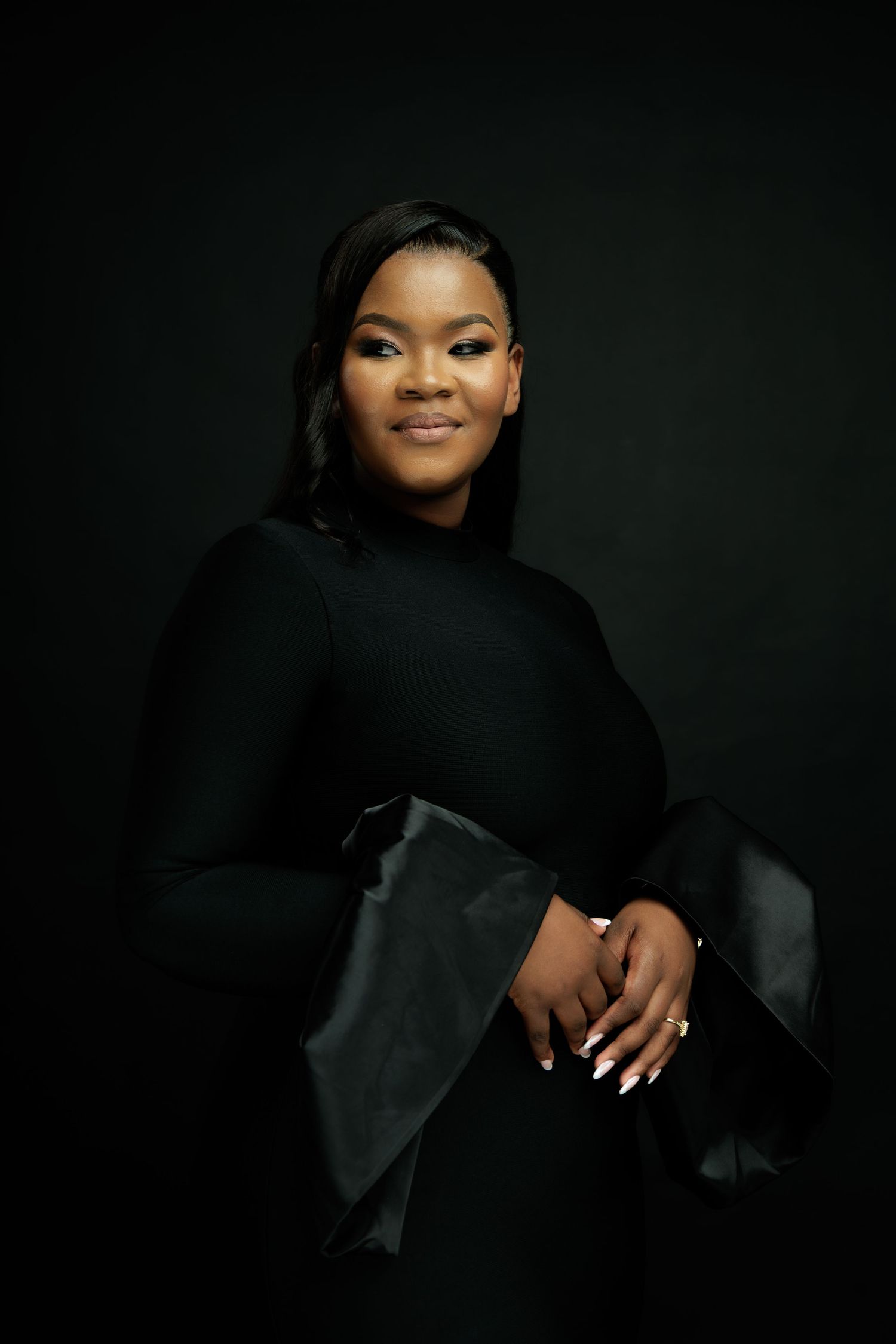 Elegant woman in black dress poses gracefully against dark studio background.