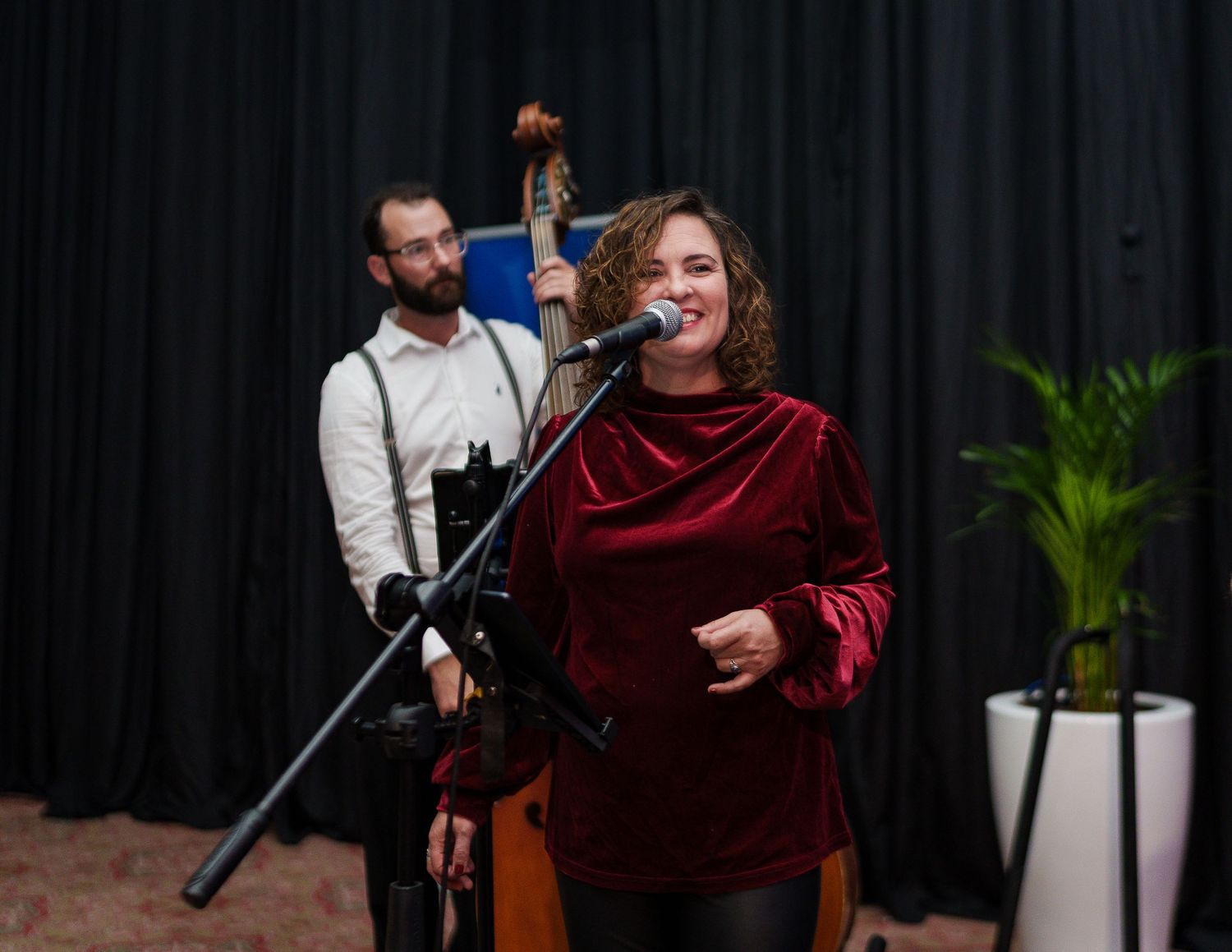Woman in red velvet top singing at microphone with bassist performing behind her at live music event.