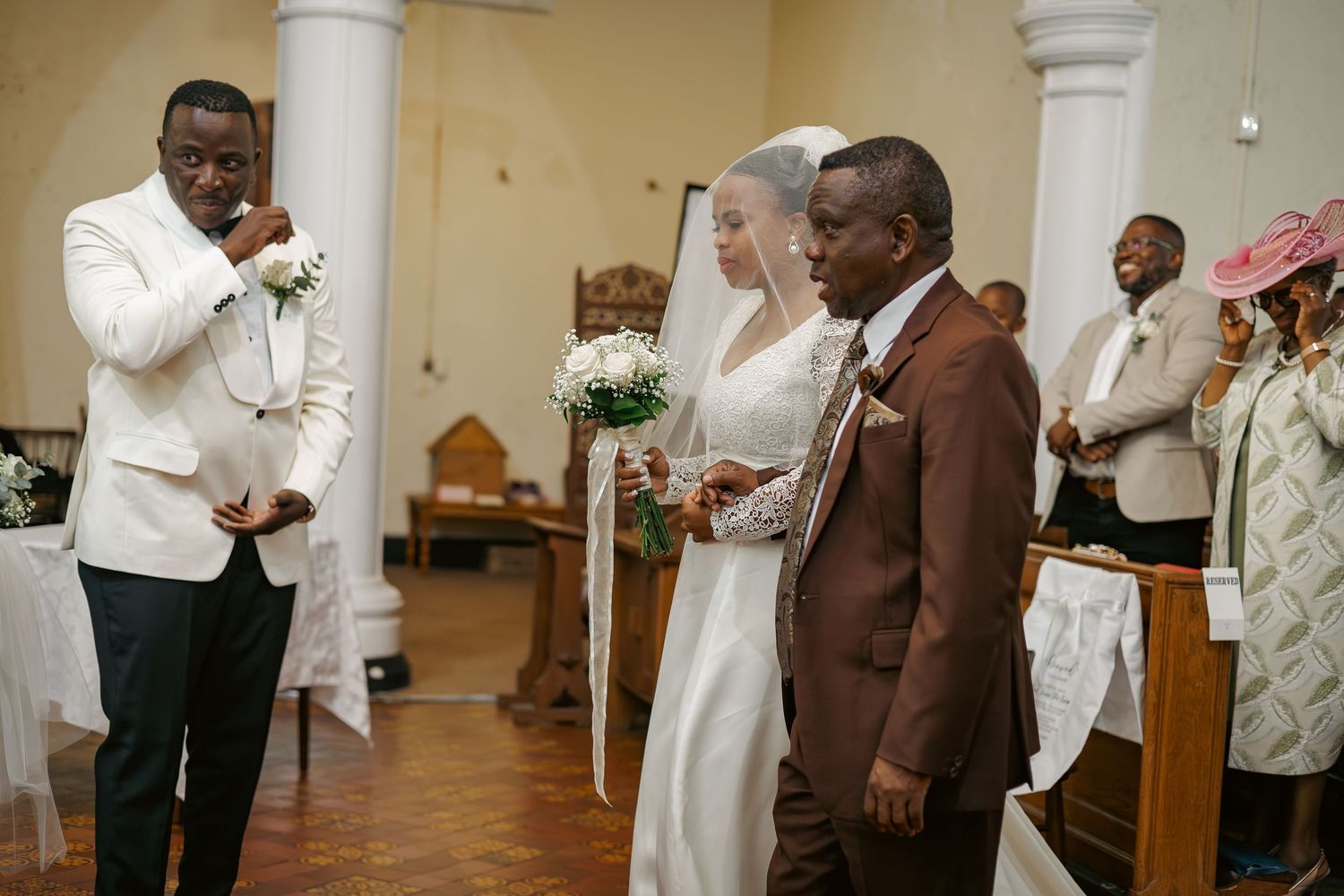 Bride in white gown escorted down church aisle by father toward groom in white suit.