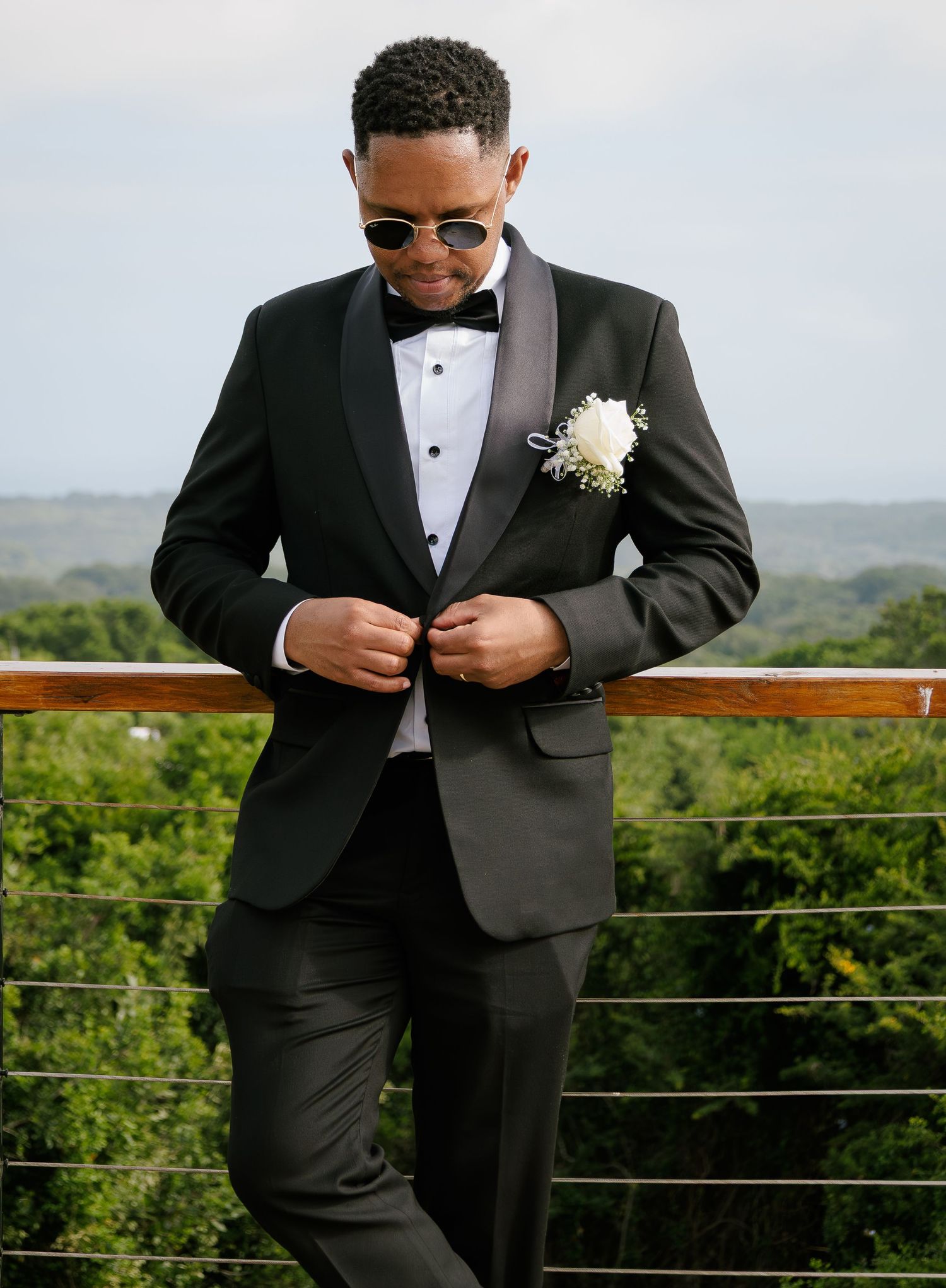 Dapper groom in black tuxedo adjusting jacket with sunglasses on a wooden deck with green backdrop.