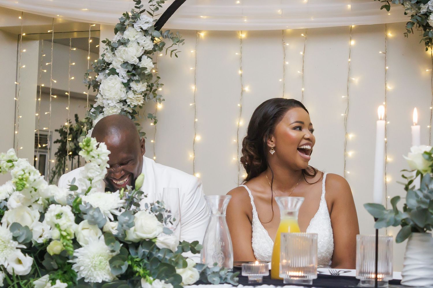 Bride and groom laugh together at the sweetheart table adorned with white flowers and fairy lights.