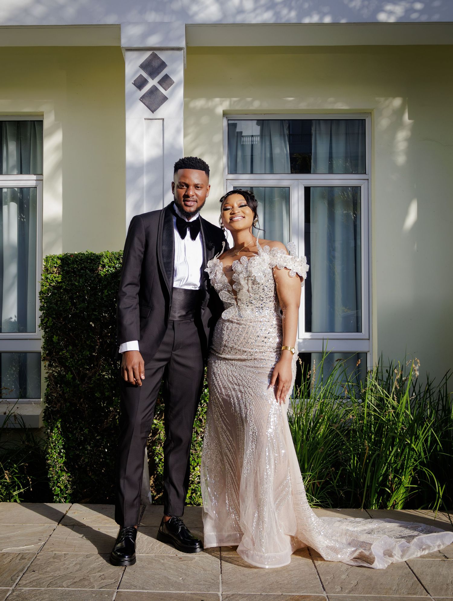 Elegant couple posing outdoors, man in black tuxedo and woman in silver beaded mermaid gown.