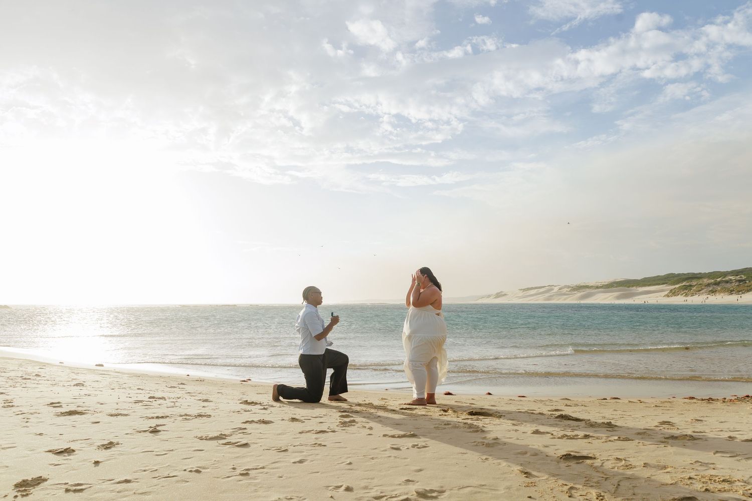 Romantic beach proposal at sunset with a man on one knee before a woman in a white dress.